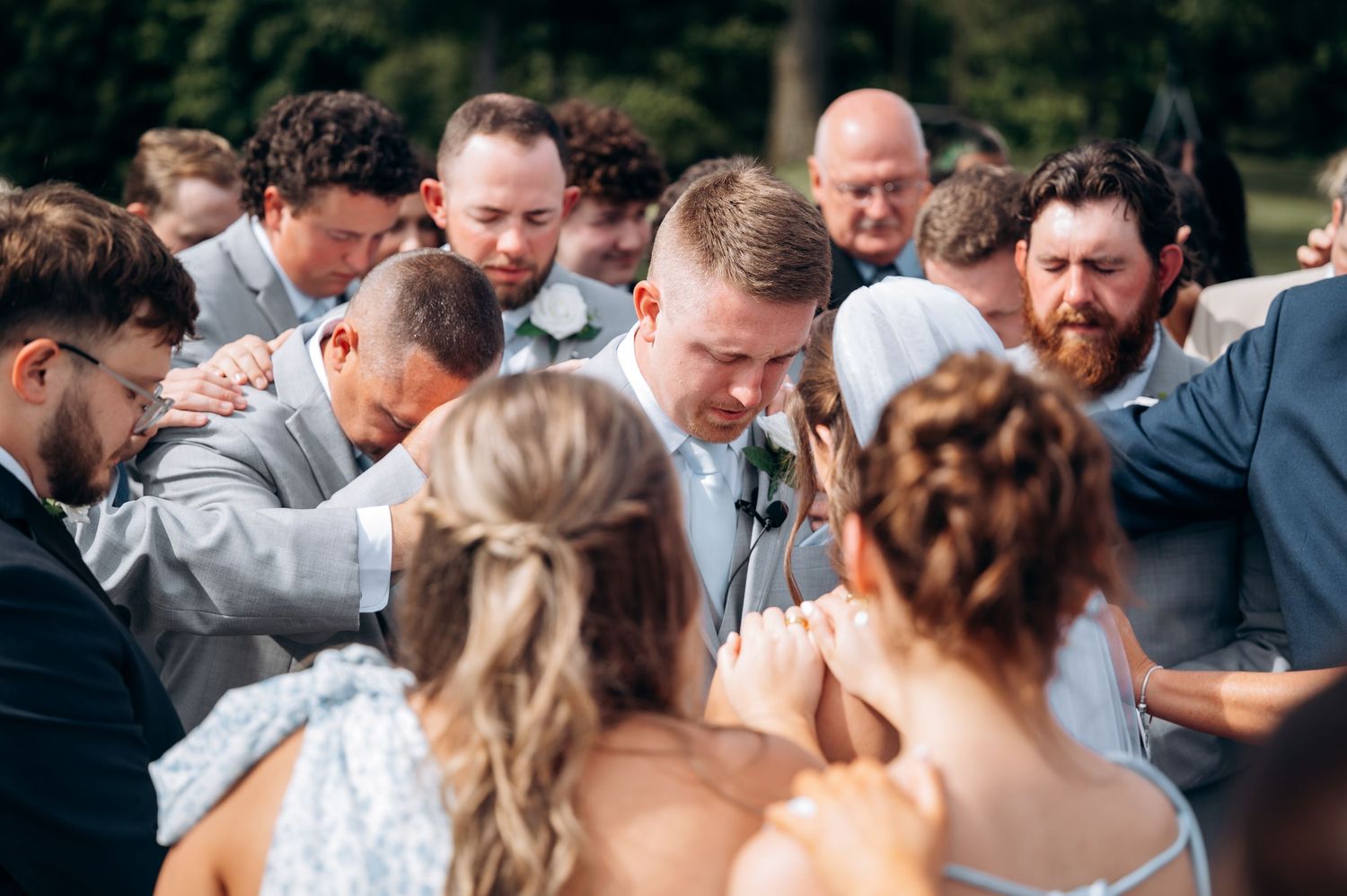 Wedding guests gather in a circle, placing hands on the groom in a heartfelt prayer moment outdoors.