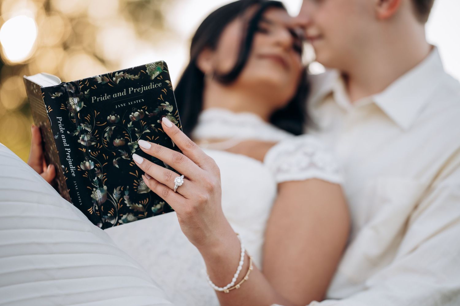 Couple sharing a romantic moment while reading Pride and Prejudice book outdoors, woman wearing engagement ring.