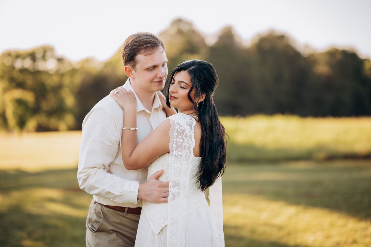 Couple in romantic embrace outdoors at golden hour, bride in white lace dress and groom in cream shirt on green field.
