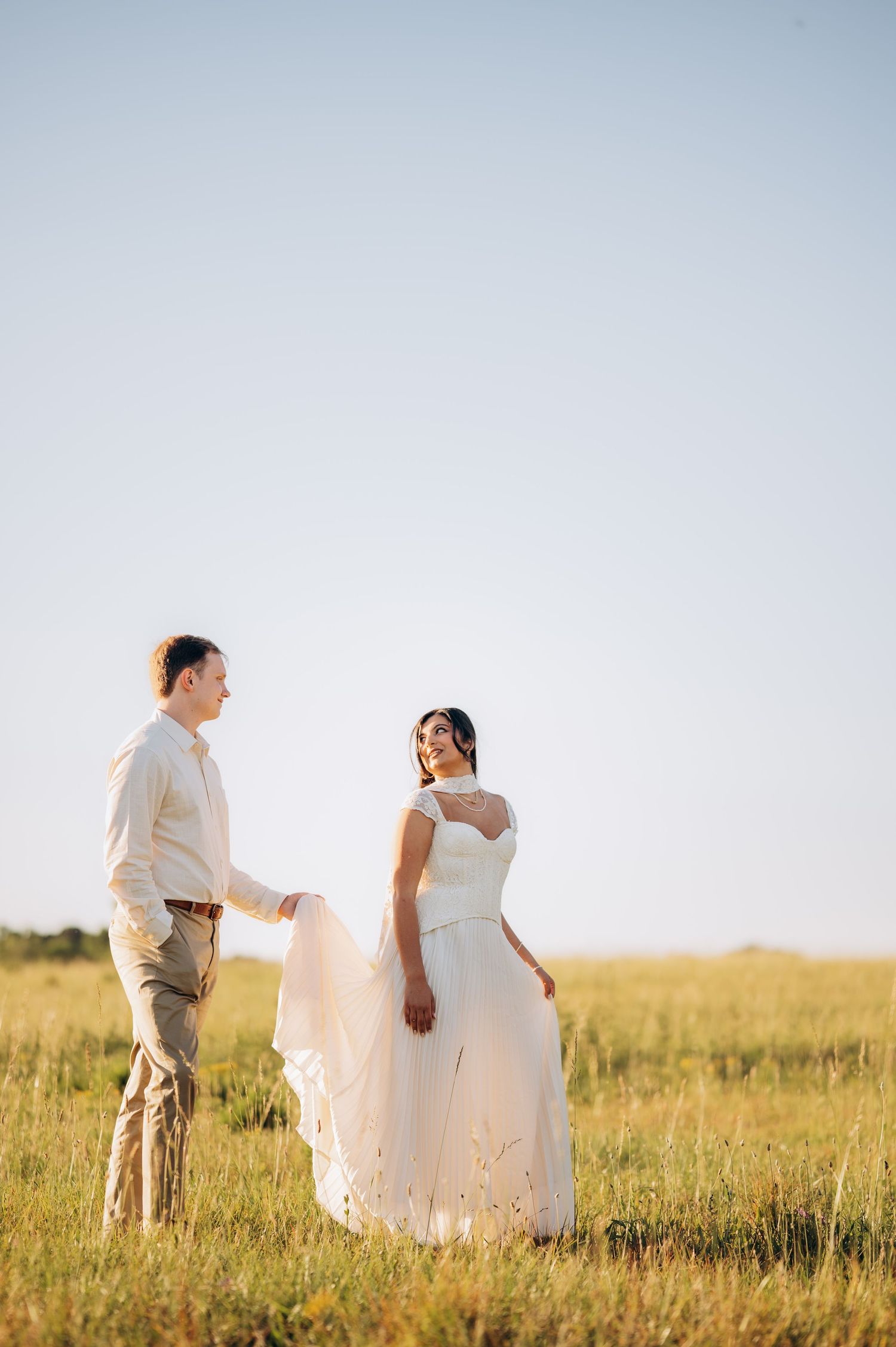Bride and groom share a romantic moment in a sunlit open field, with the bride's gown flowing in the breeze.