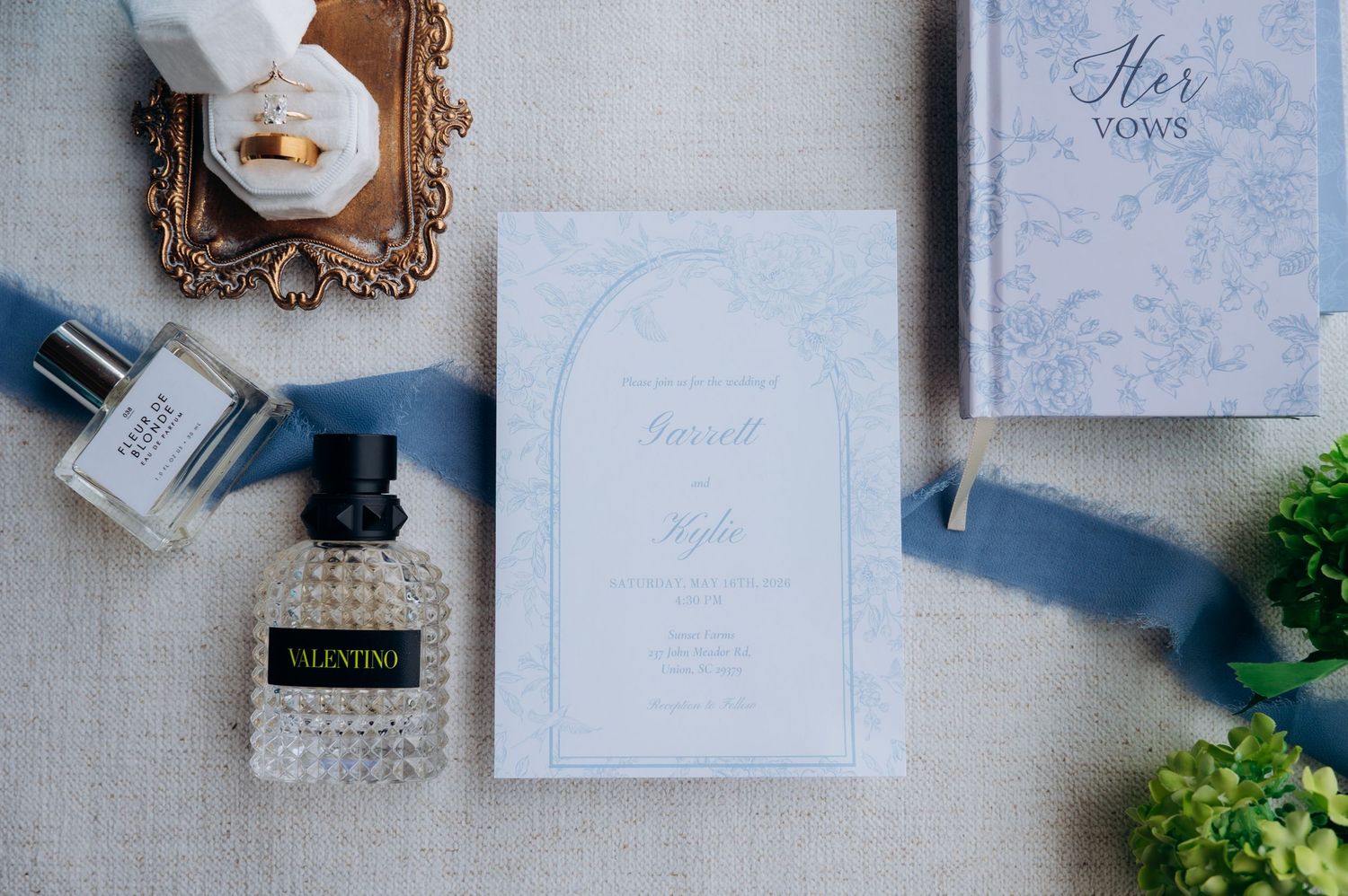 Elegant wedding flat lay with blue invitation suite, Valentino perfume, vow book, and silk ribbon on linen.