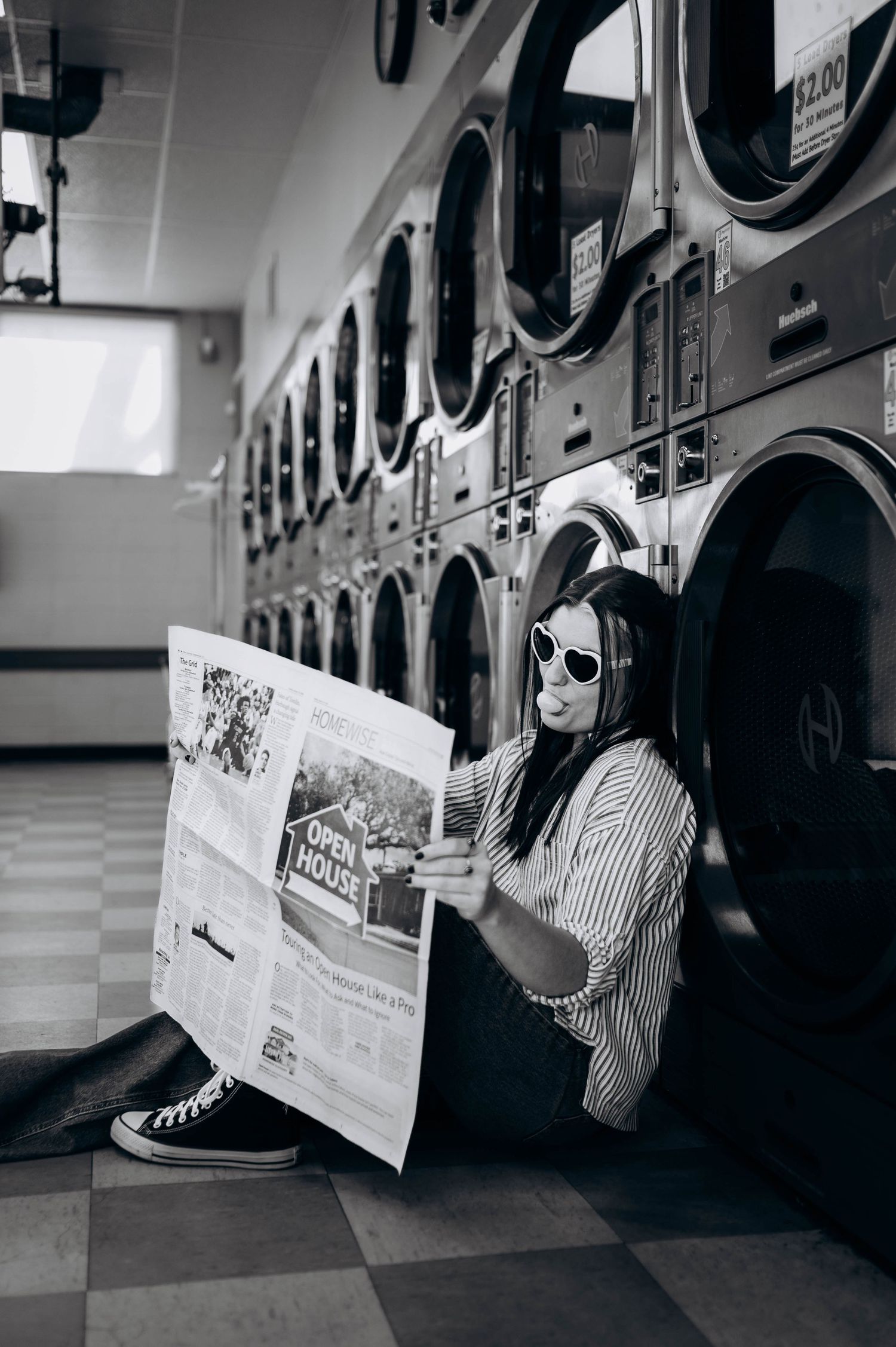Person reading newspaper while sitting on floor of laundromat with row of washing machines in background.