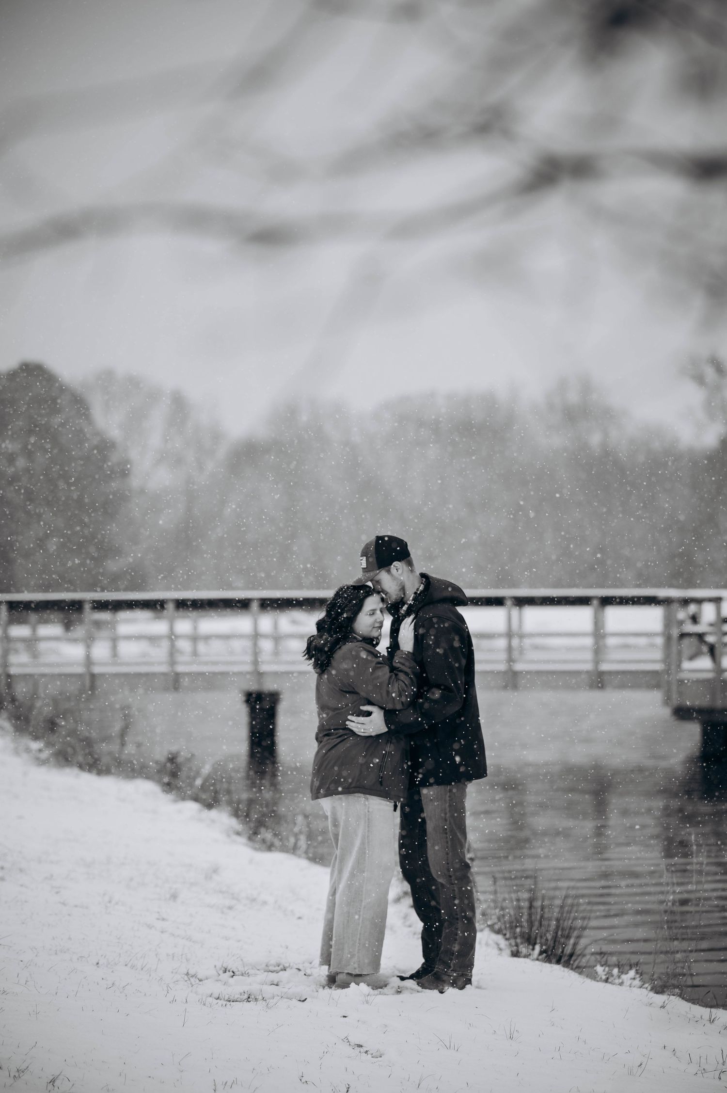 Two figures embrace in a romantic winter scene as snow falls gently around them by a bridge.