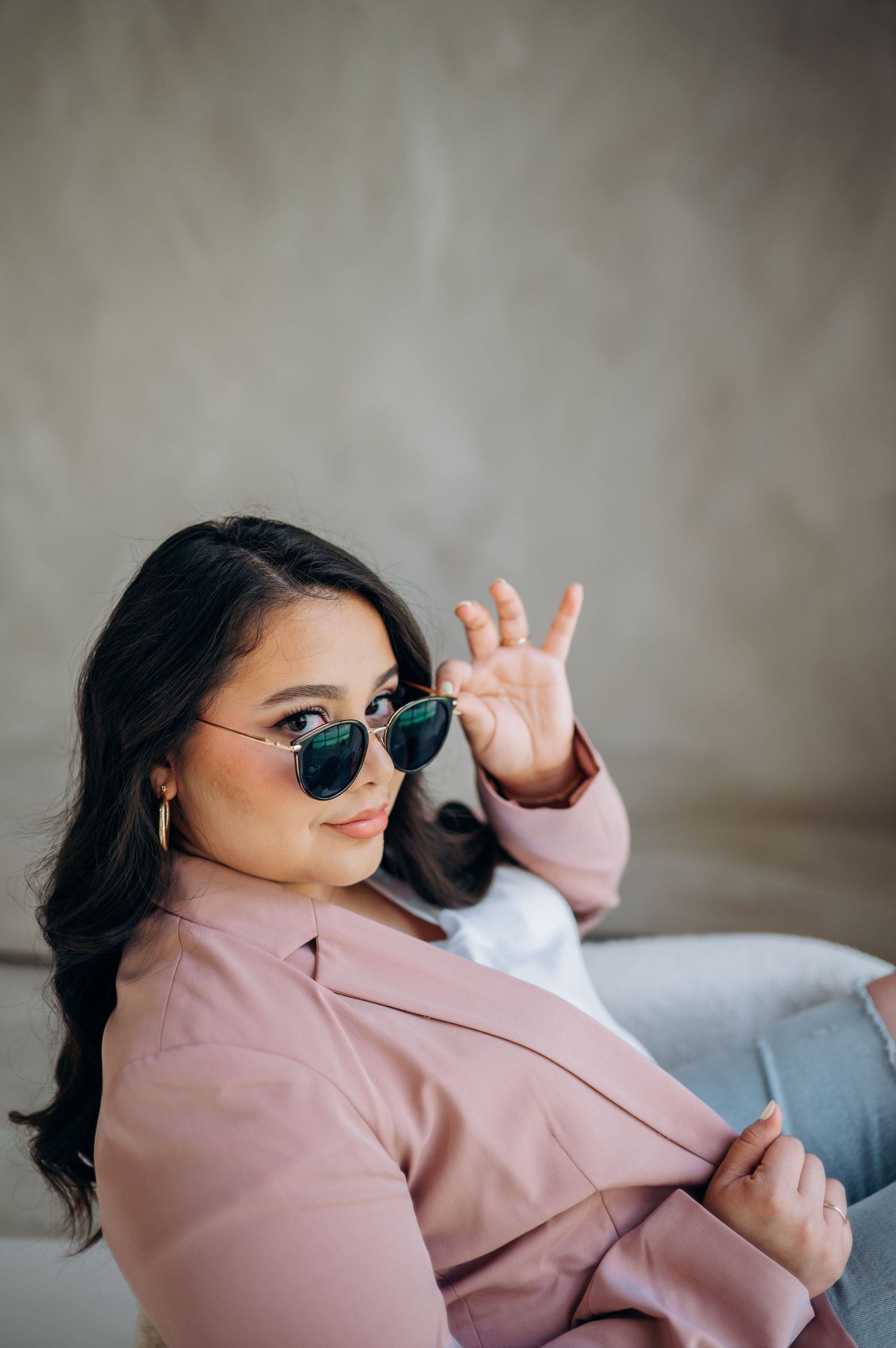Fashionable portrait with sunglasses and pink blazer against textured wall background.