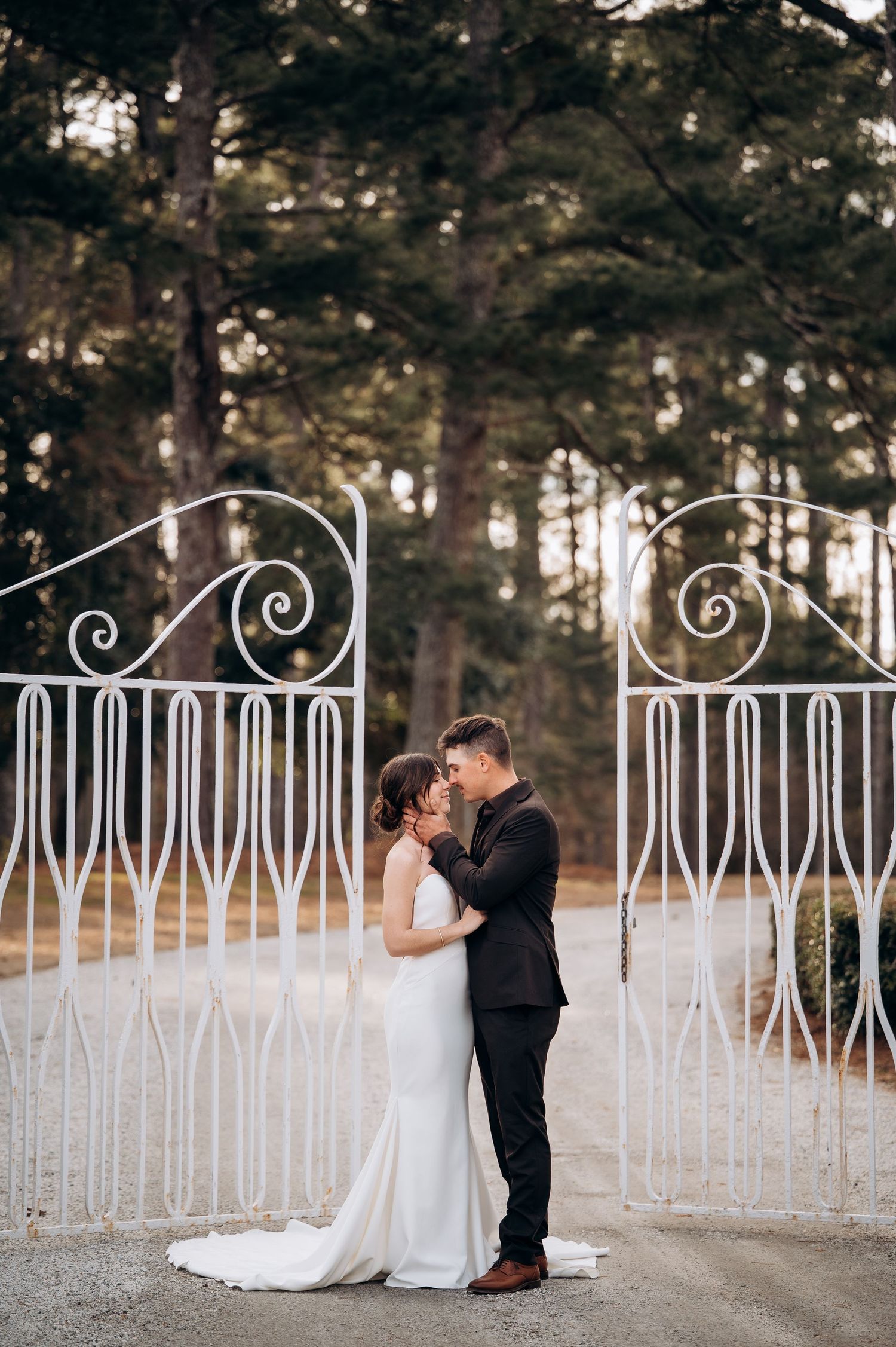 A romantic wedding couple embraces in front of an ornate white iron gate with pine trees in the background.