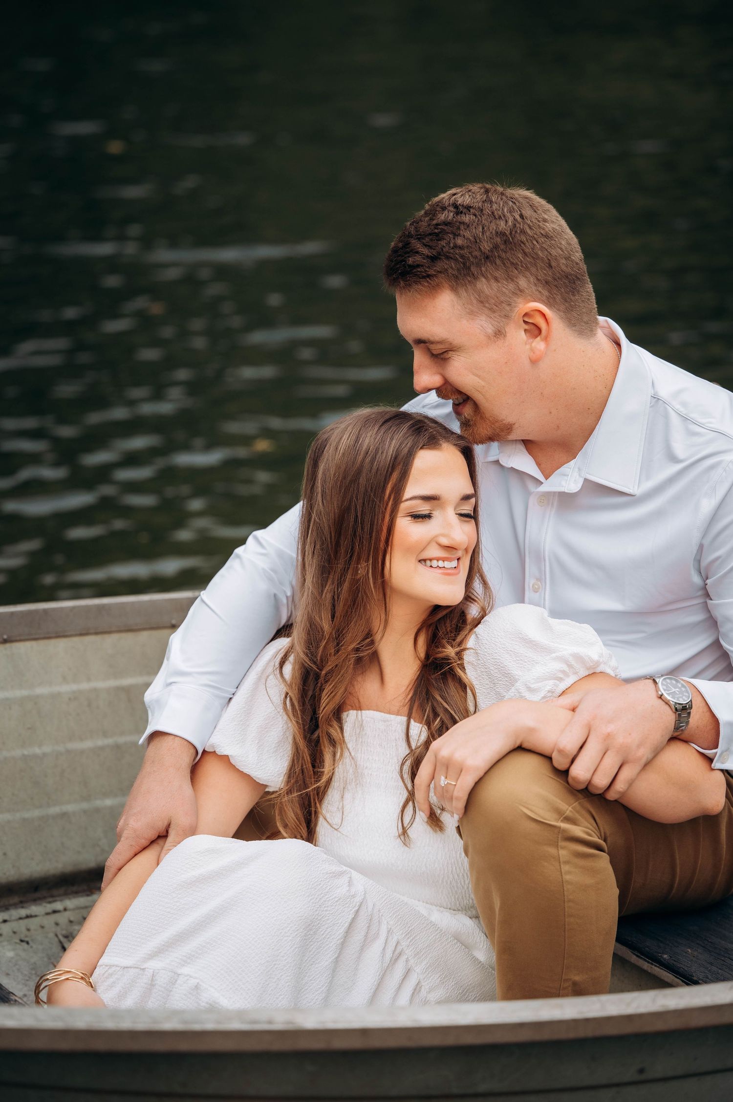 A couple relaxes in a rowboat on a lake wearing white and khaki casual outfits.