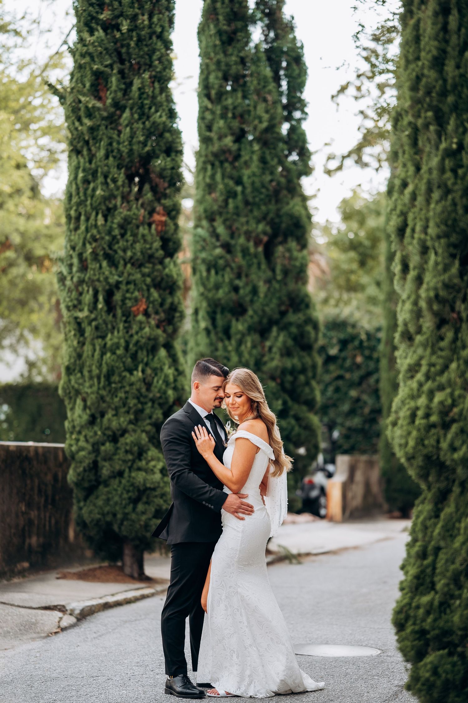 A bride and groom embrace romantically on a tree-lined path, with tall cypress trees in the background.