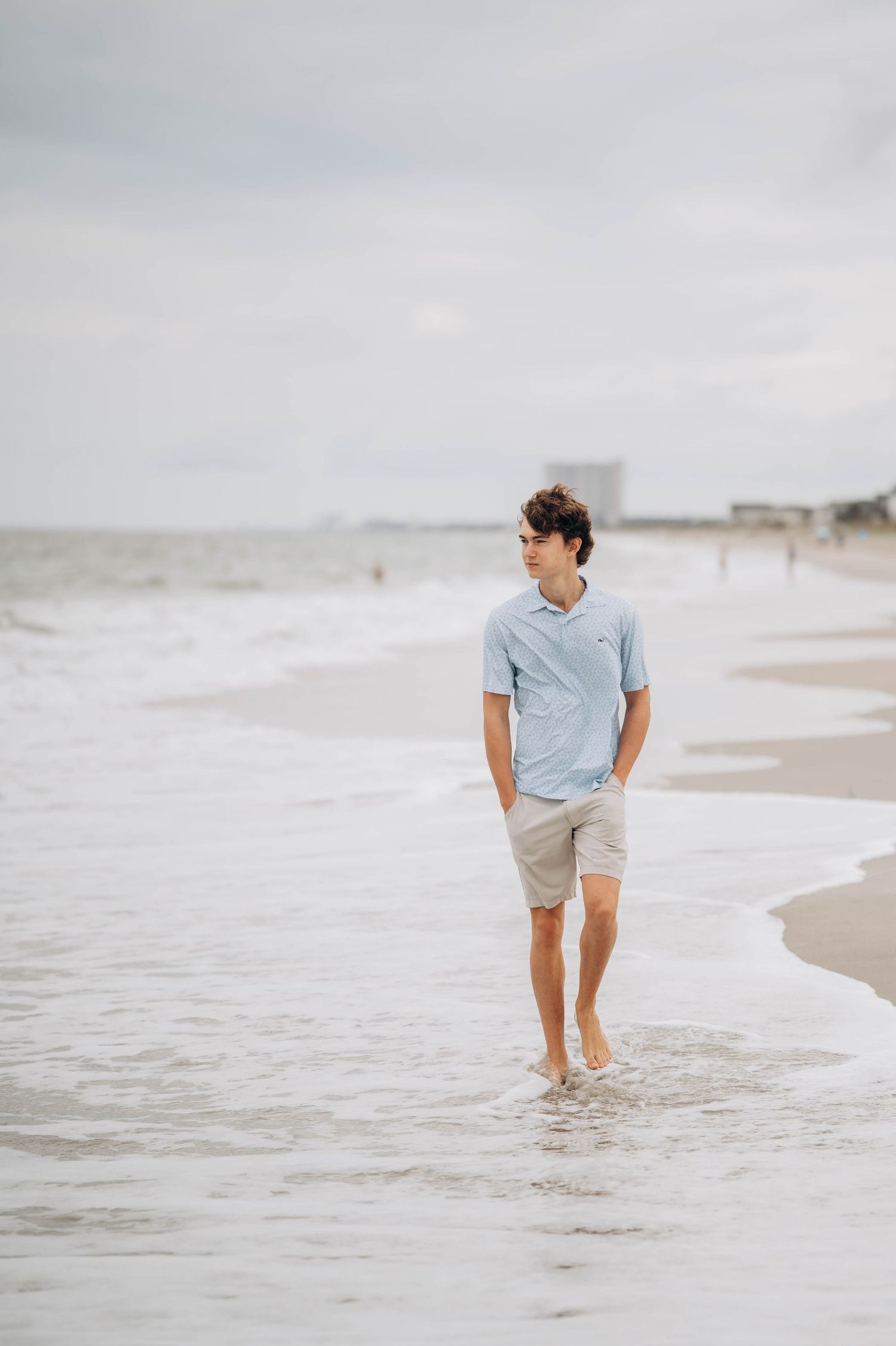 Person in light blue shirt walks along ocean shoreline on cloudy day.