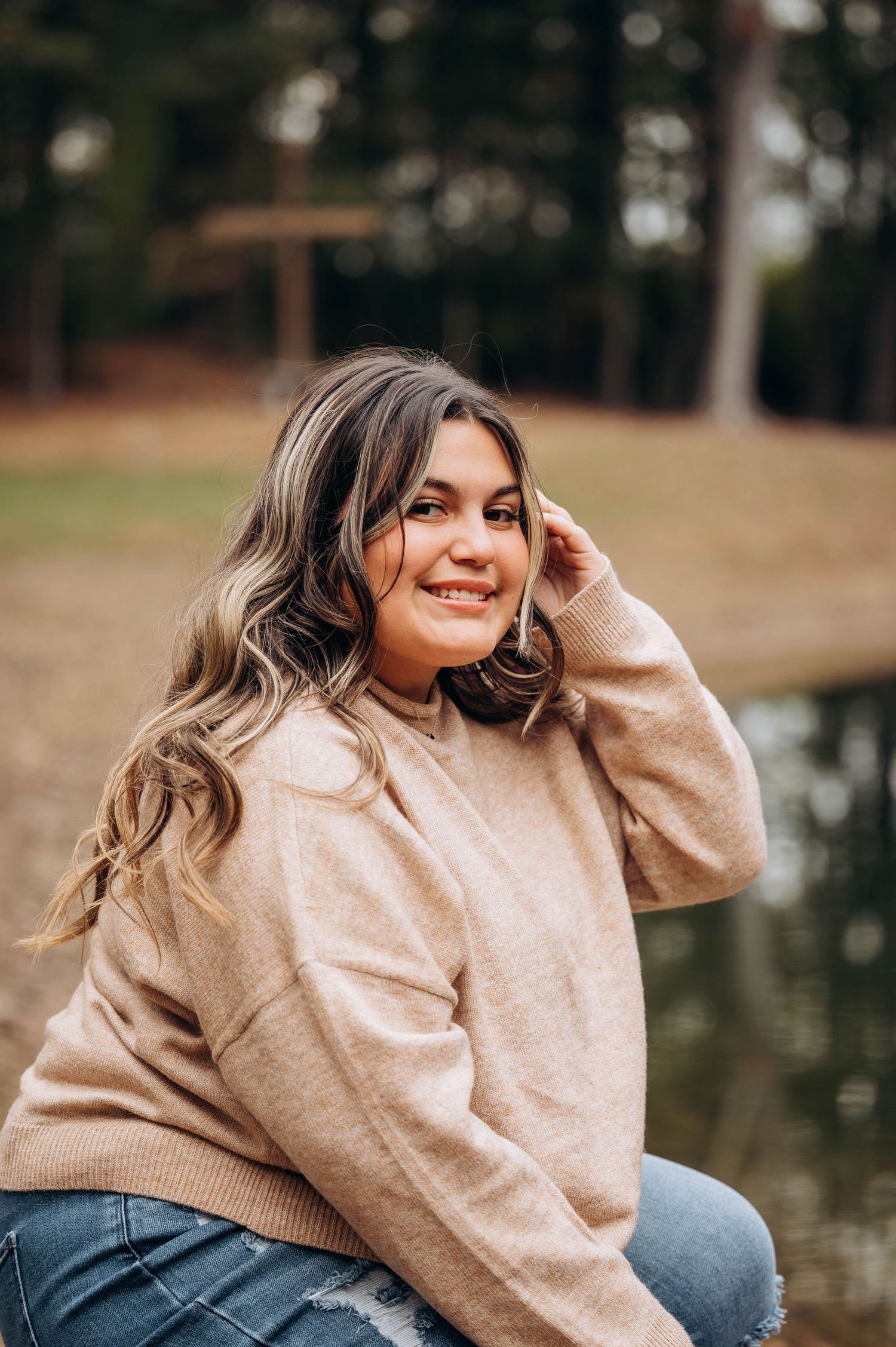 Woman in beige sweater smiles while sitting outdoors near a lake.