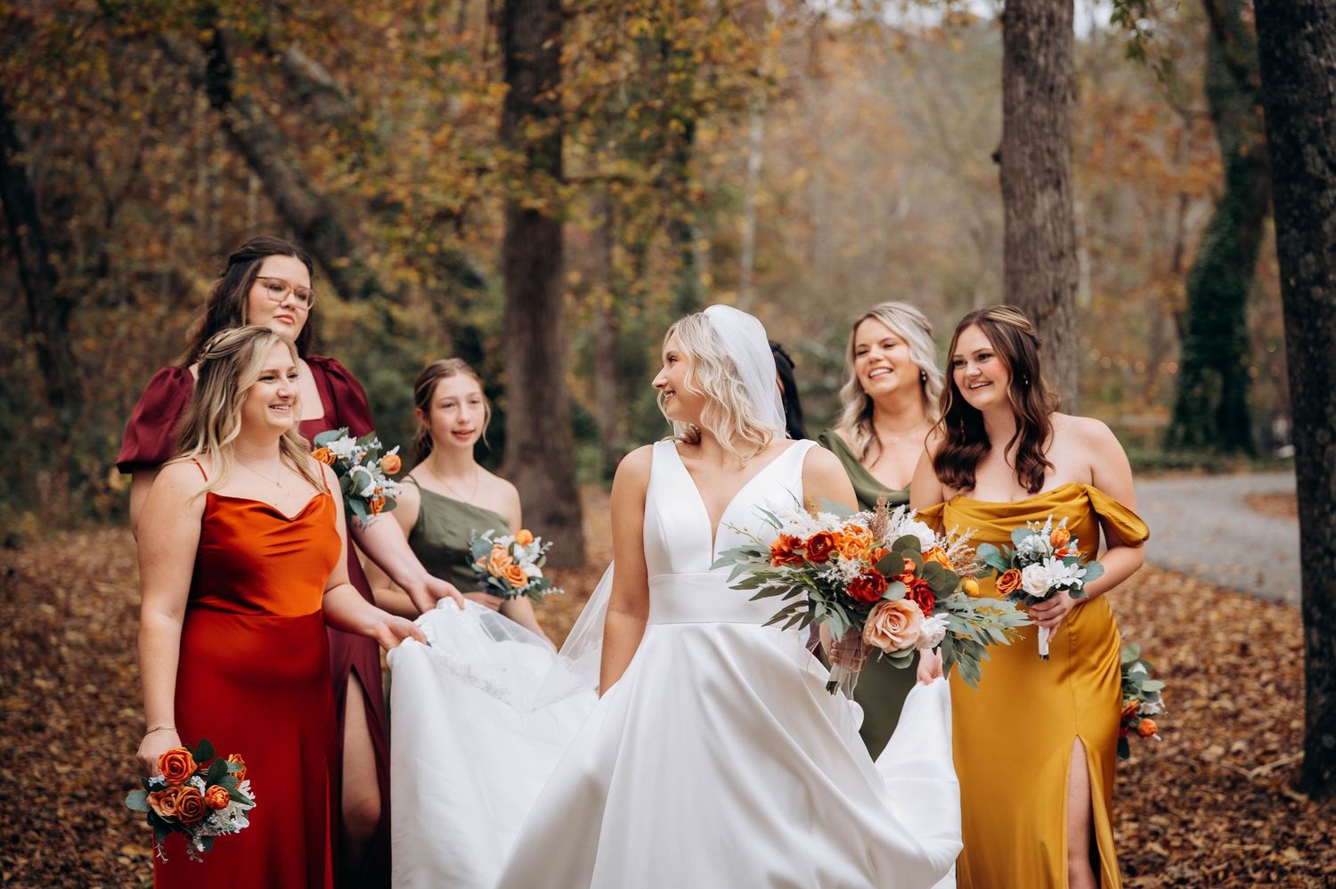 Bridal party wearing burgundy and mustard dresses standing together in an autumn forest setting.