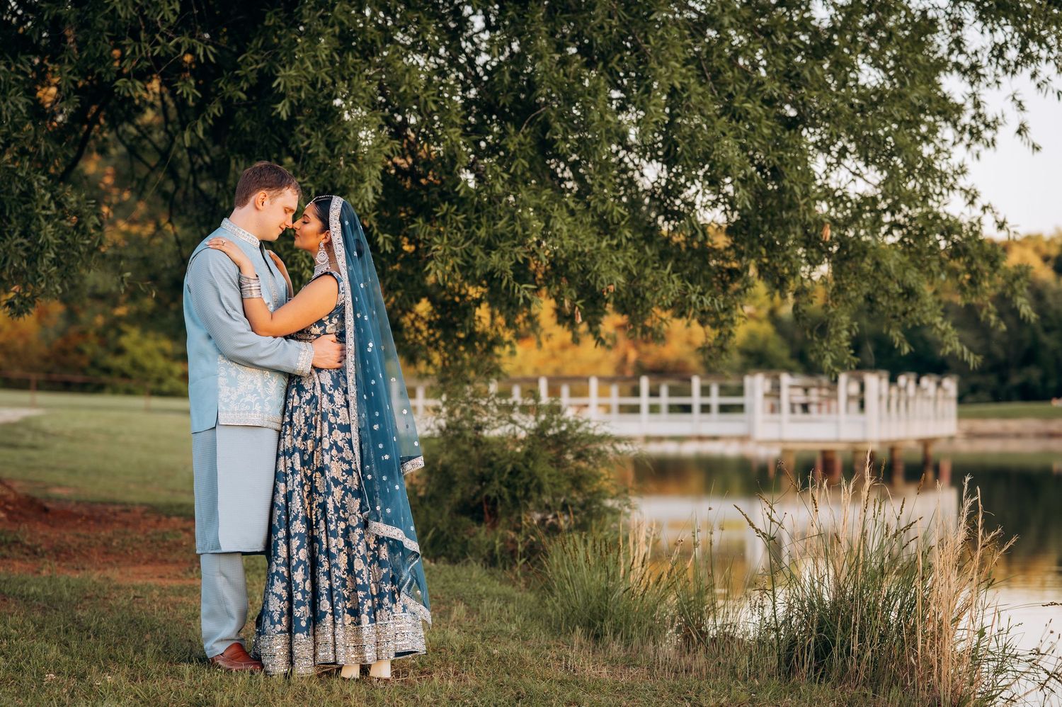 Couple in wedding attire embracing under a tree by a serene pond at golden hour outdoor ceremony.