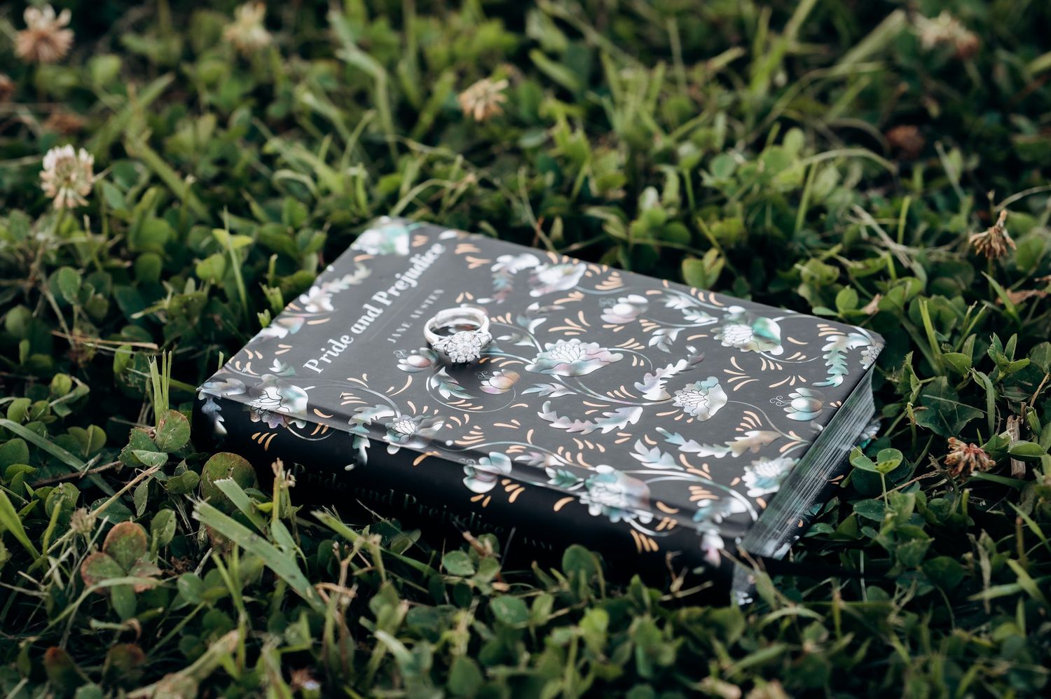 A floral-patterned Pride and Prejudice book with a ring resting on top, lying in lush green grass.