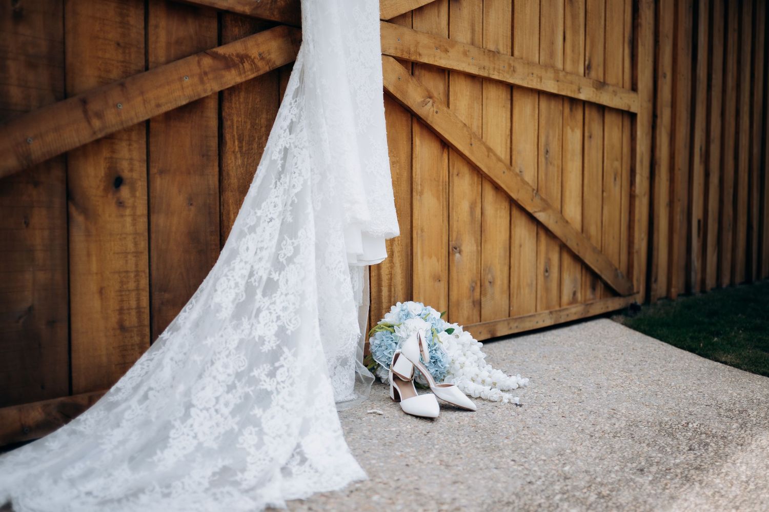 Elegant lace wedding dress train flowing beside bridal bouquet and white heels against rustic wooden barn doors.