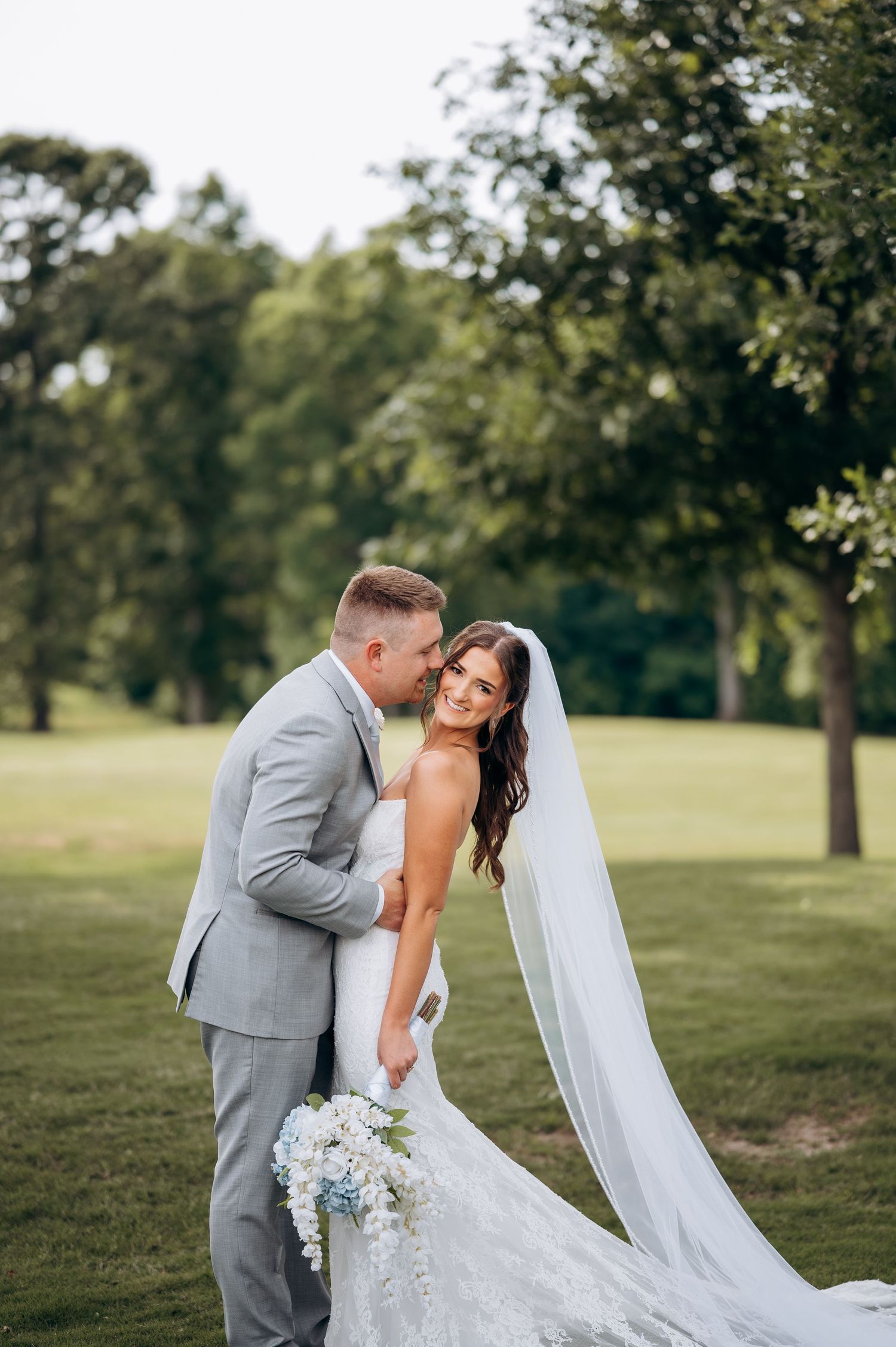 Smiling bride in lace gown with cathedral veil holds blue bouquet as groom kisses her cheek outdoors.