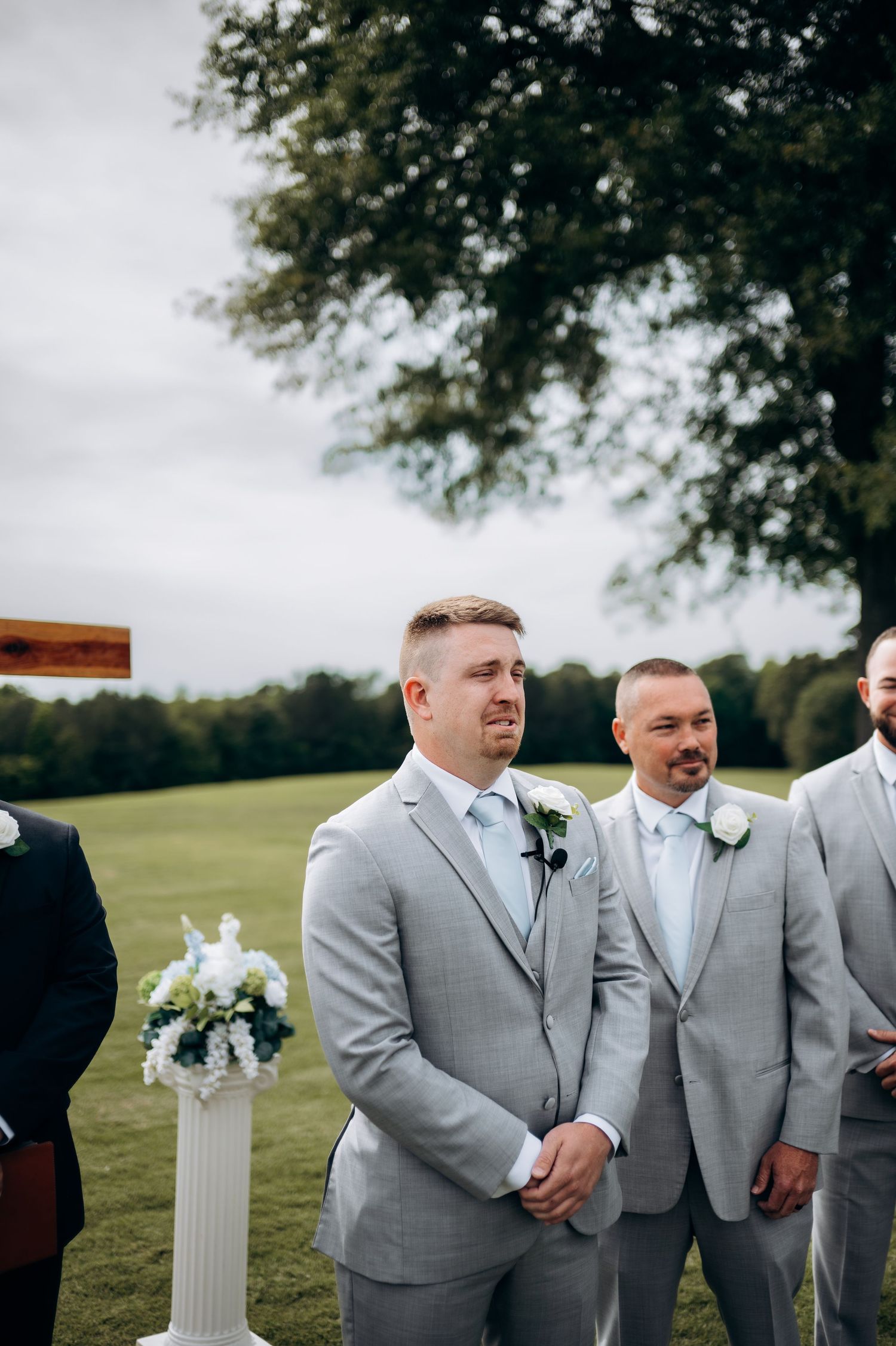 Emotional groom in gray suit watches bride walk down the aisle at outdoor wedding ceremony.