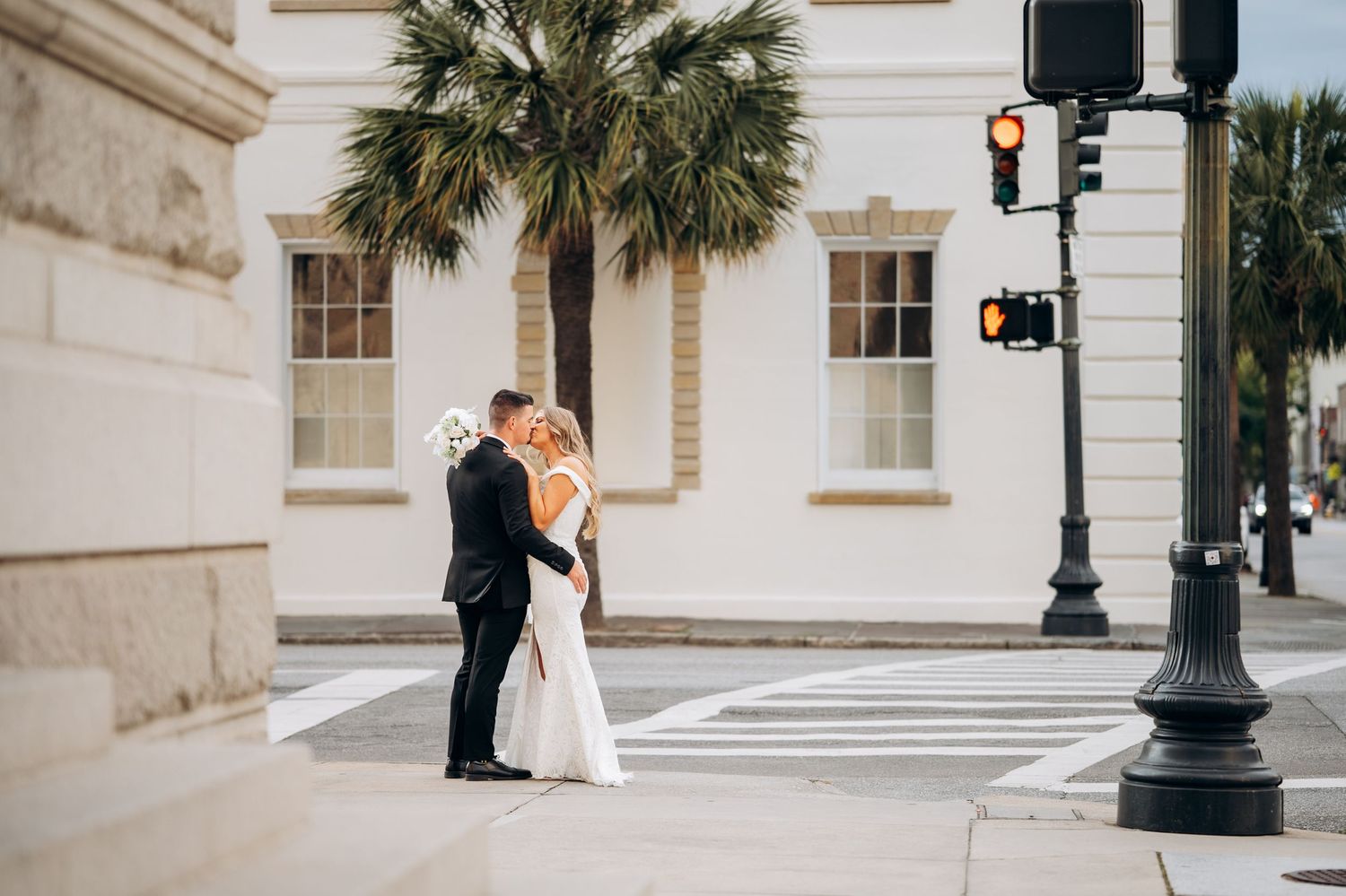 Bride and groom share a kiss at a crosswalk in a charming downtown setting with a palm tree backdrop.