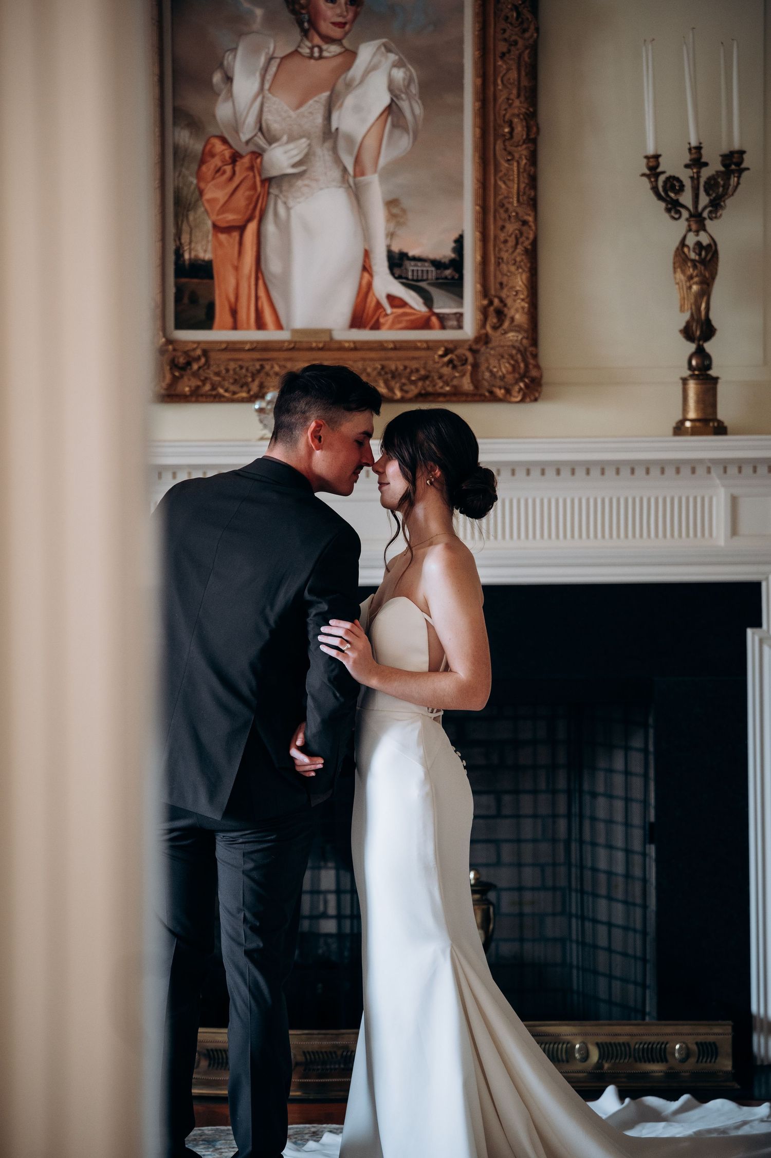 Couple sharing romantic moment before fireplace with ornate portrait painting and candelabra in elegant room.