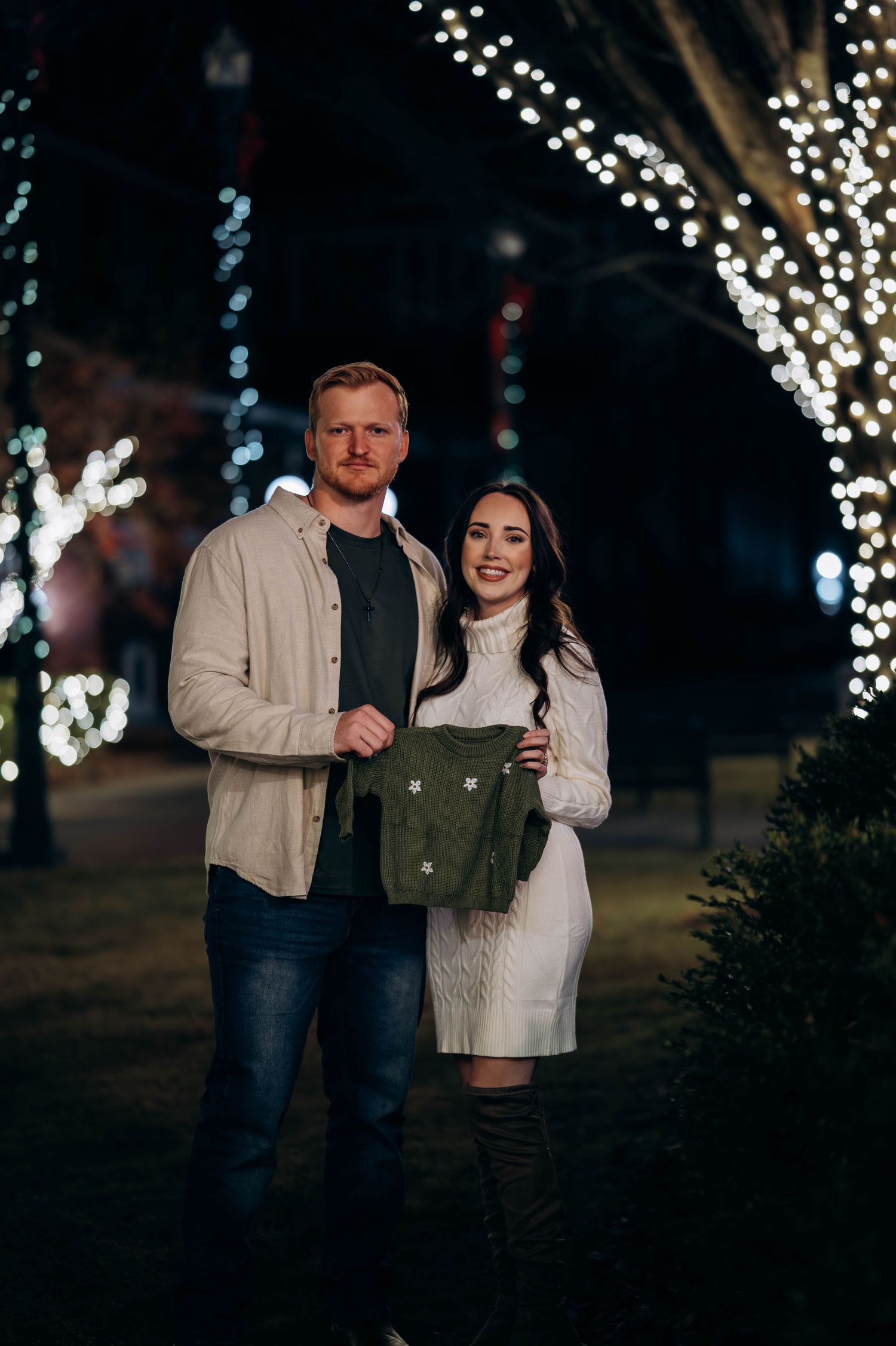 Couple posing outdoors at night holding a green baby outfit surrounded by festive string lights.