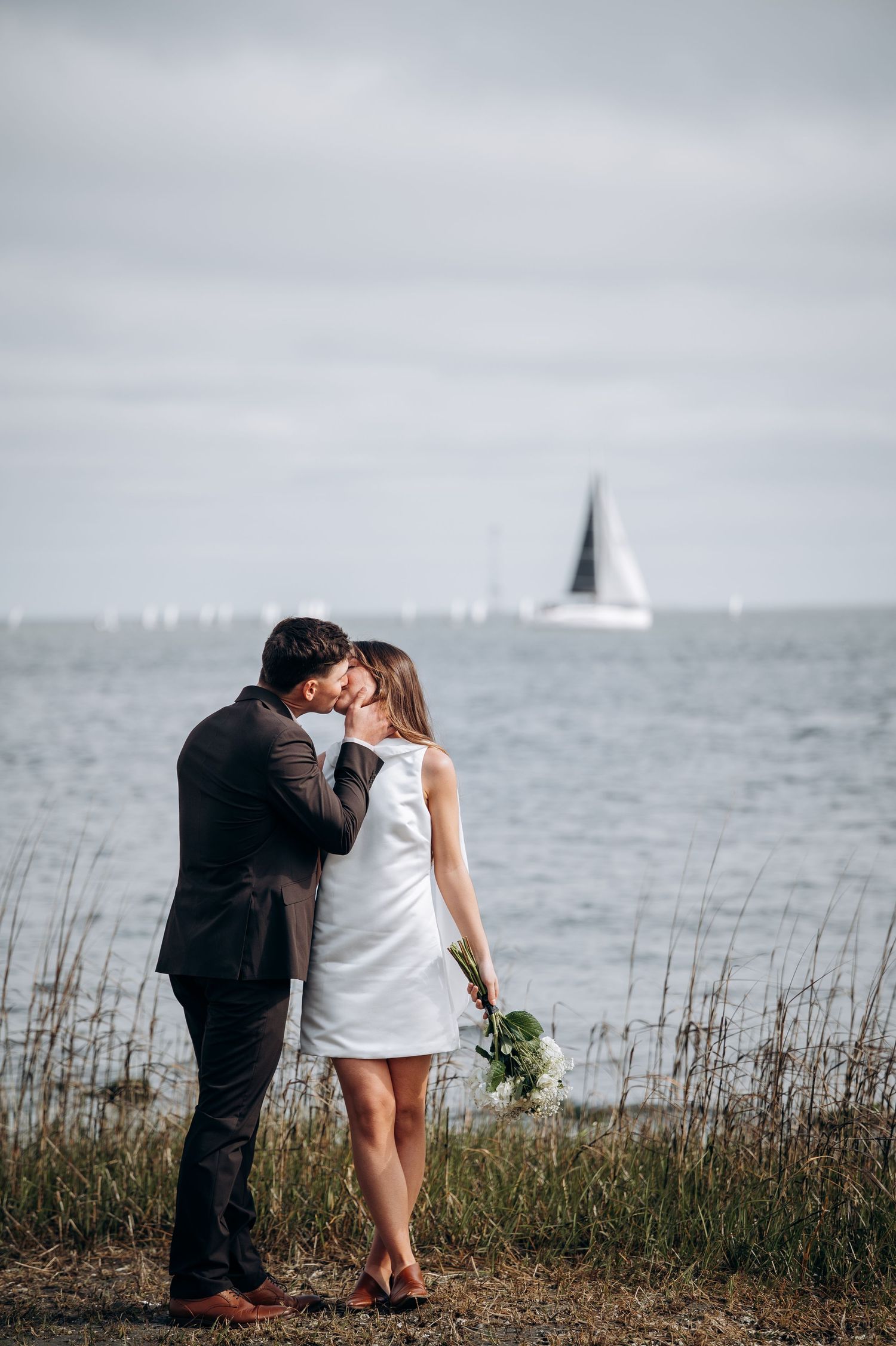 Couple shares a kiss by the waterfront, bride holding a white bouquet with a sailboat visible in the background.