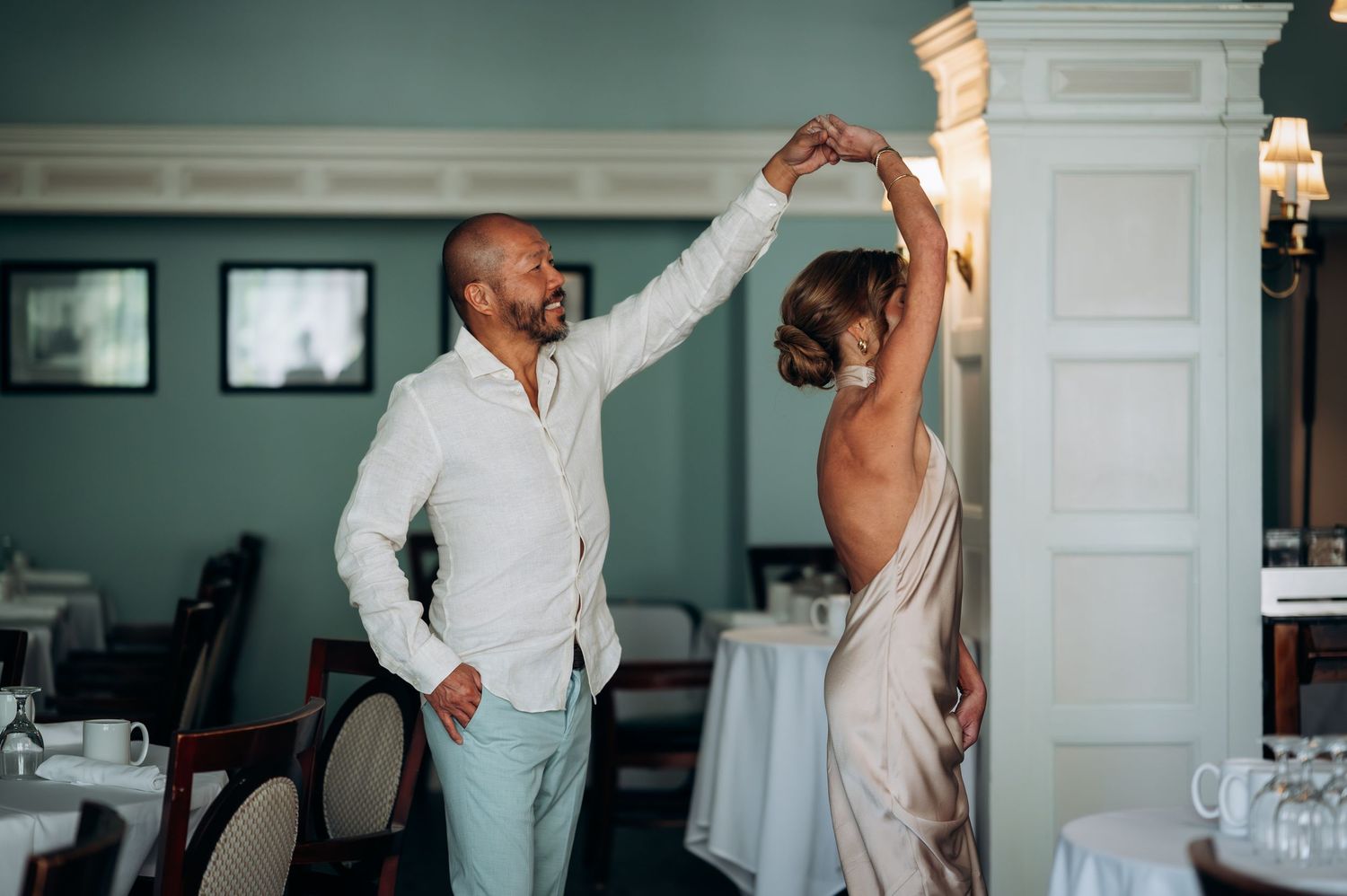 Couple sharing a romantic first dance in an elegant teal dining room, groom in white shirt spinning bride in backless gown.