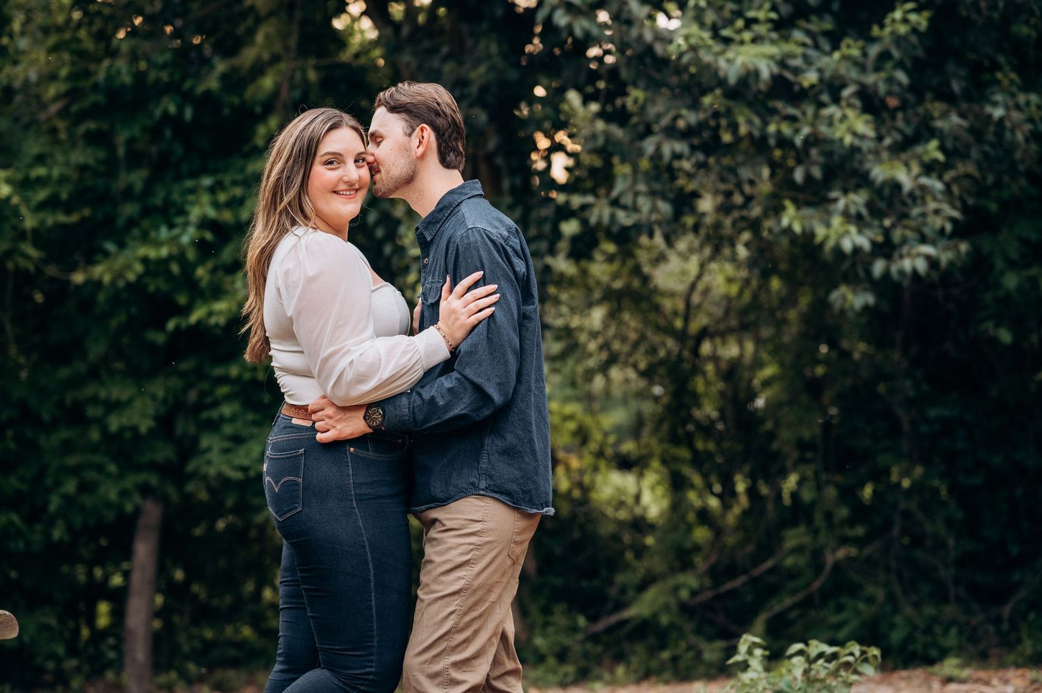 Couple embracing and smiling together in front of lush green trees at golden hour.