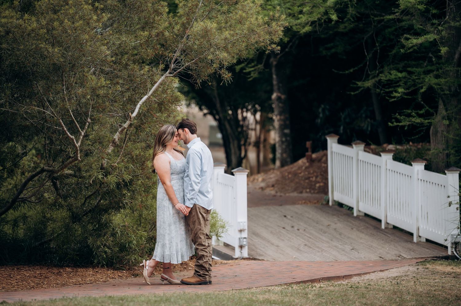 Romantic couple sharing an intimate moment on a brick path beside a white fence surrounded by lush green trees.