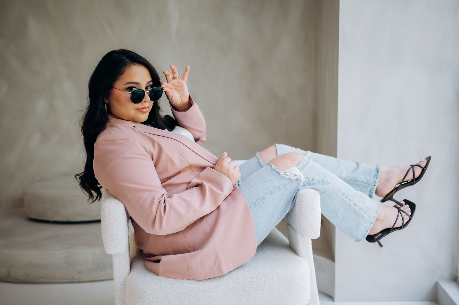 A person in a pink blazer and ripped jeans reclines on a white chair while making a peace sign gesture.