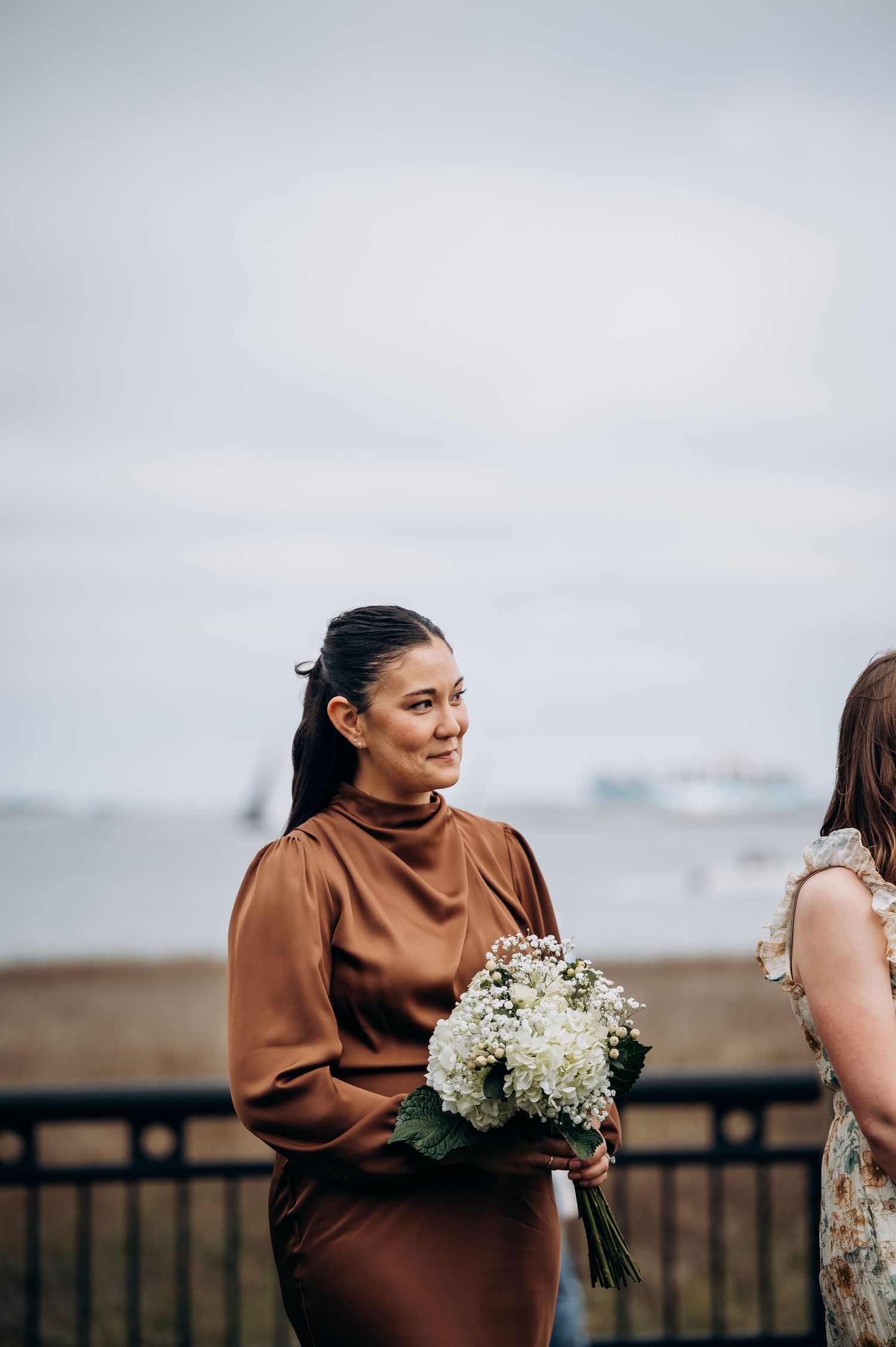 A wedding party stands together holding white floral bouquets against an overcast sky.