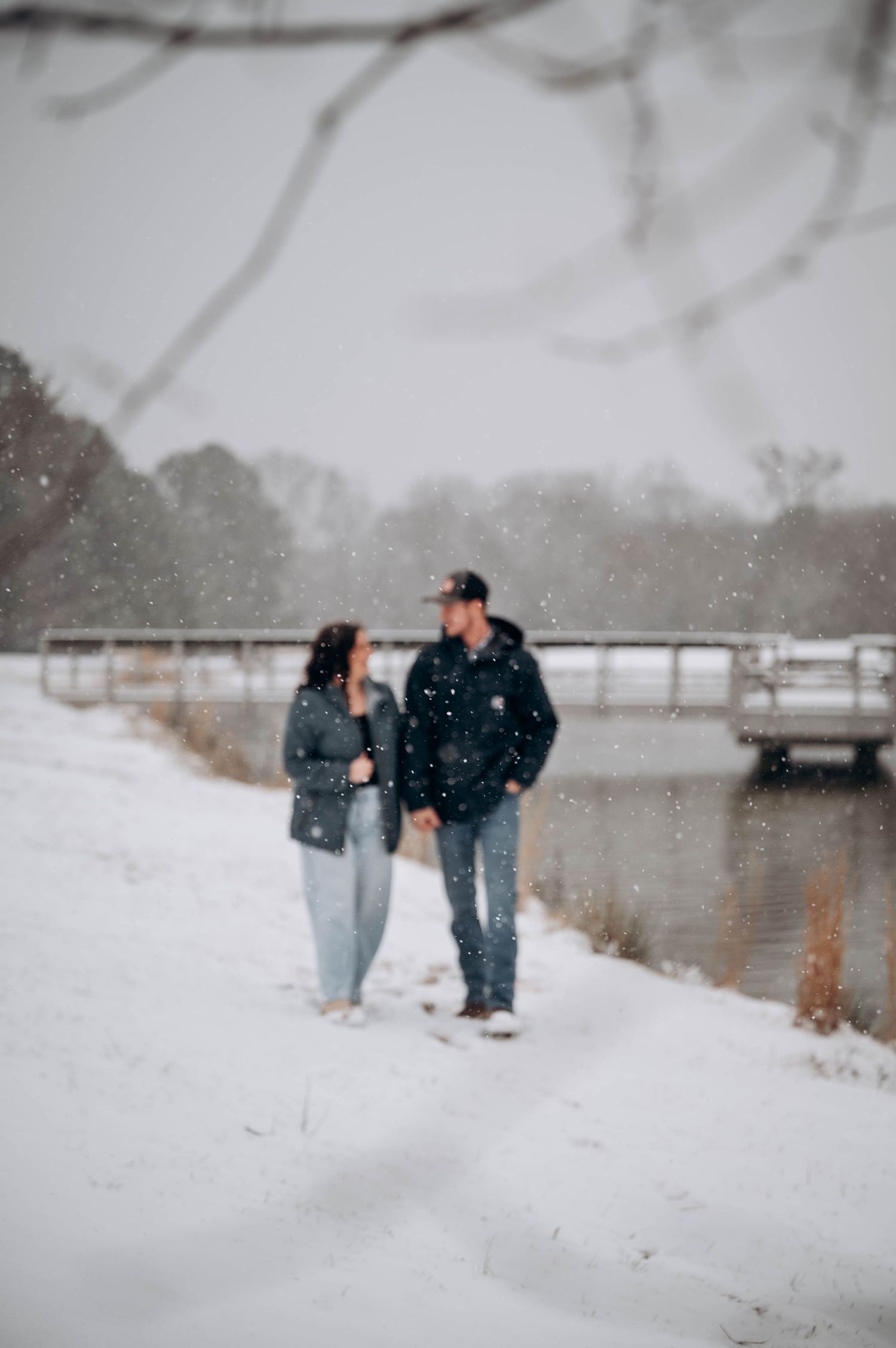 A couple walks together through a snowy winter landscape near a lake with falling snowflakes.