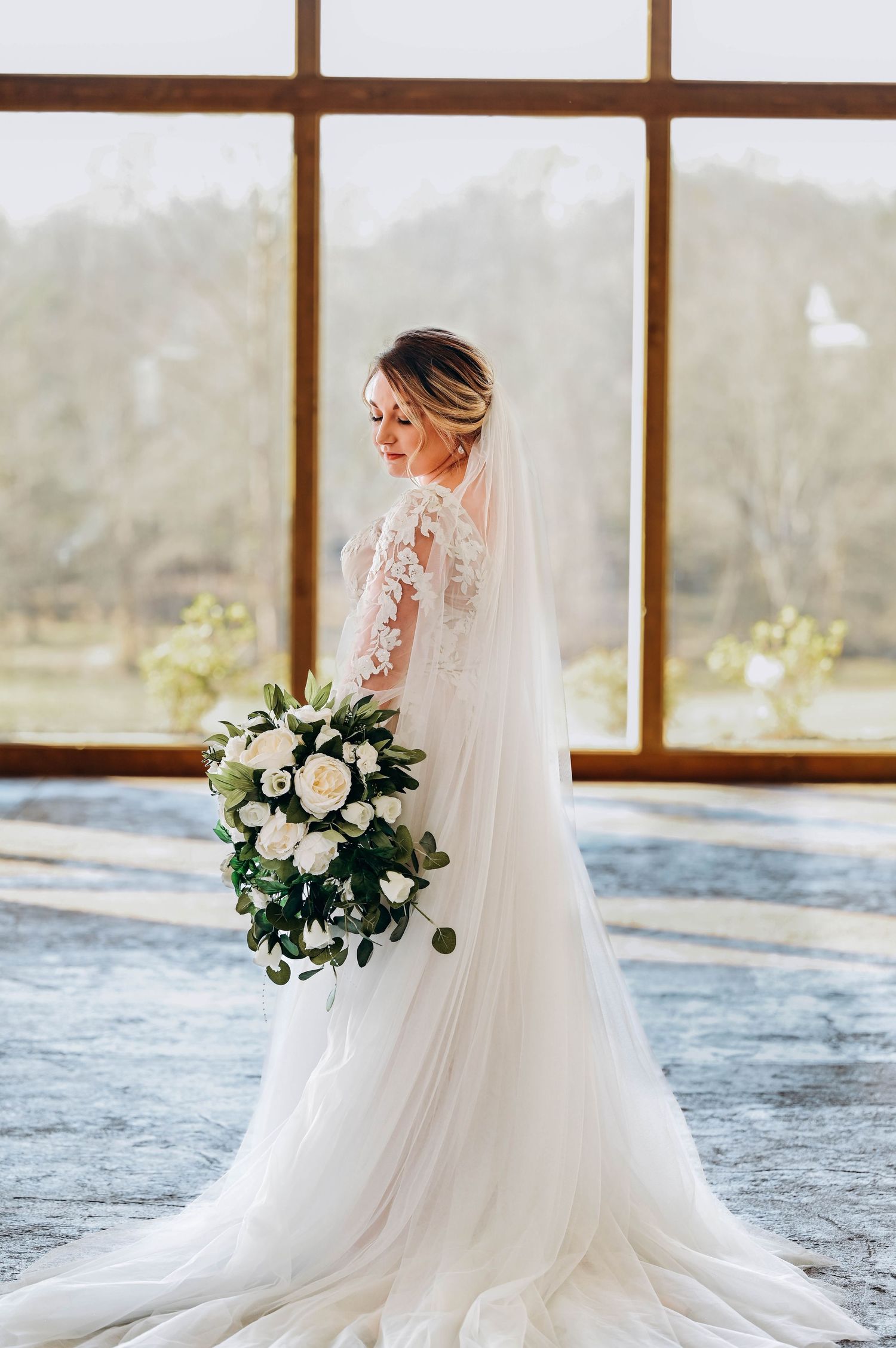 Elegant white wedding dress with flowing train and bouquet photographed against large windows in natural light.