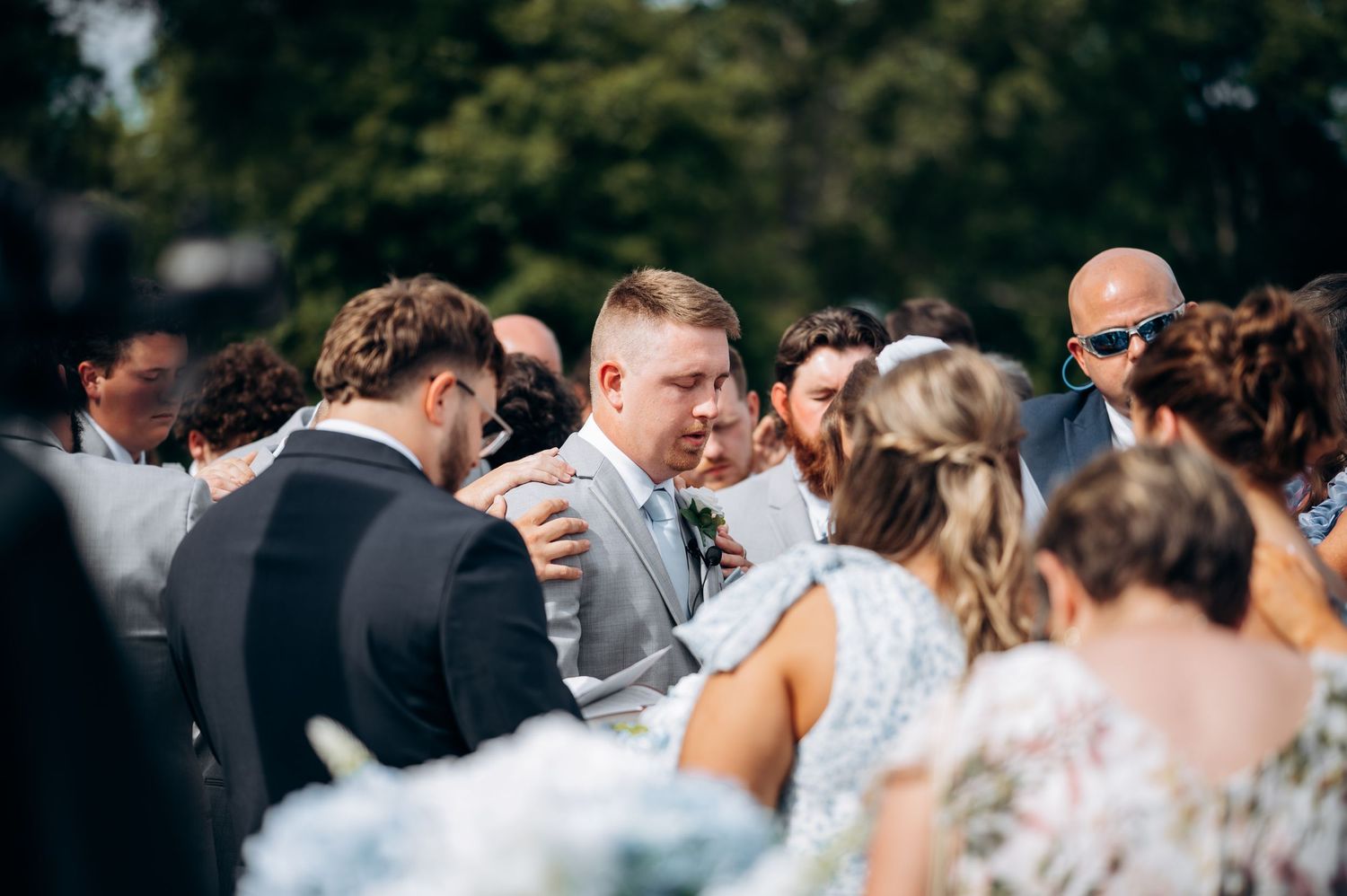 Groom embraces guests during wedding reception, surrounded by smiling attendees in an outdoor setting.