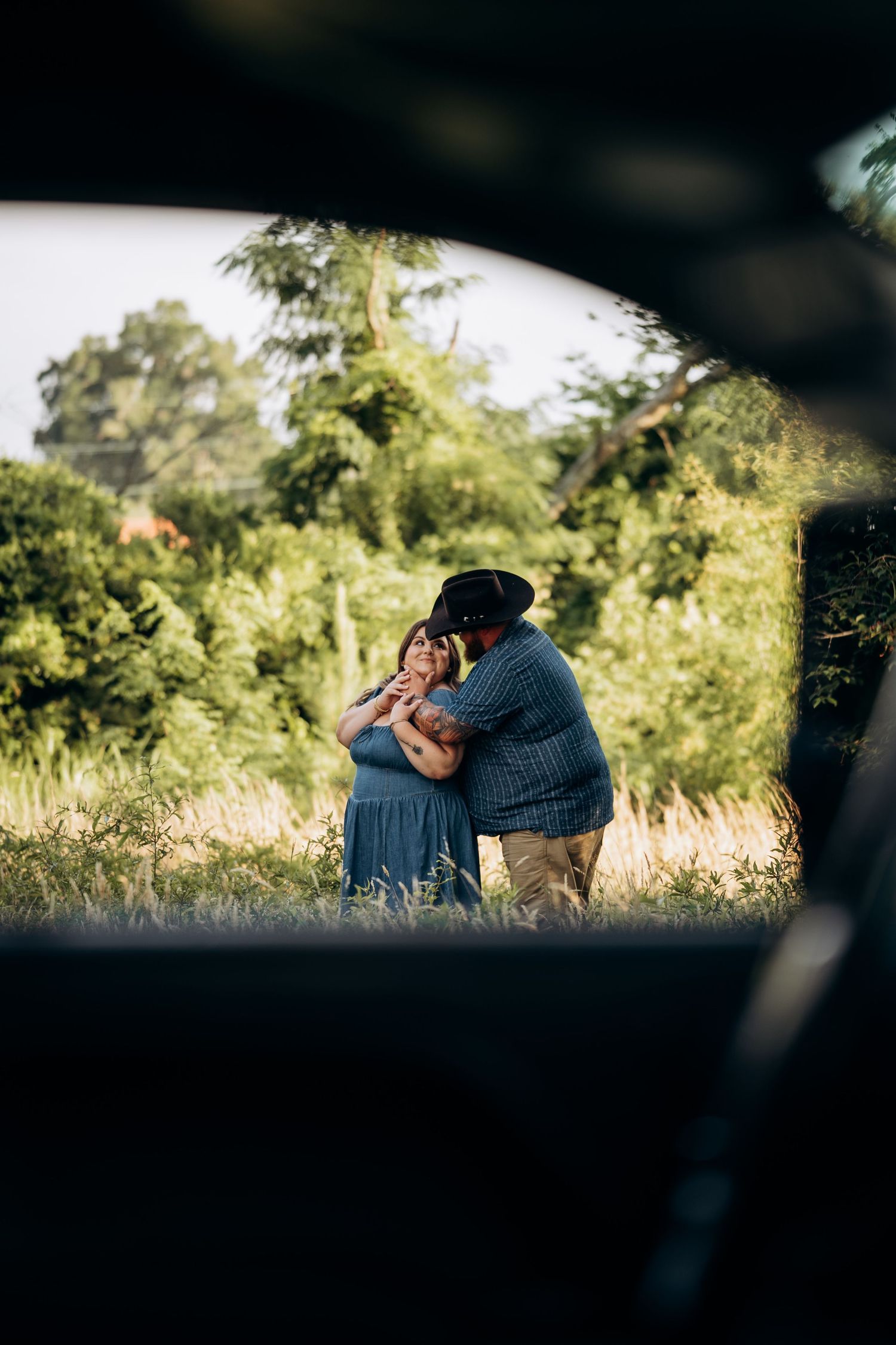 Couple embracing and kissing in a lush green field, photographed through a car window frame.