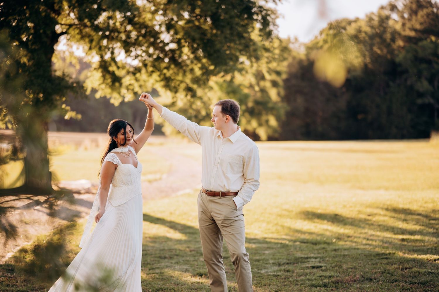 Bride and groom dancing outdoors on lush green lawn with golden sunlight filtering through trees.