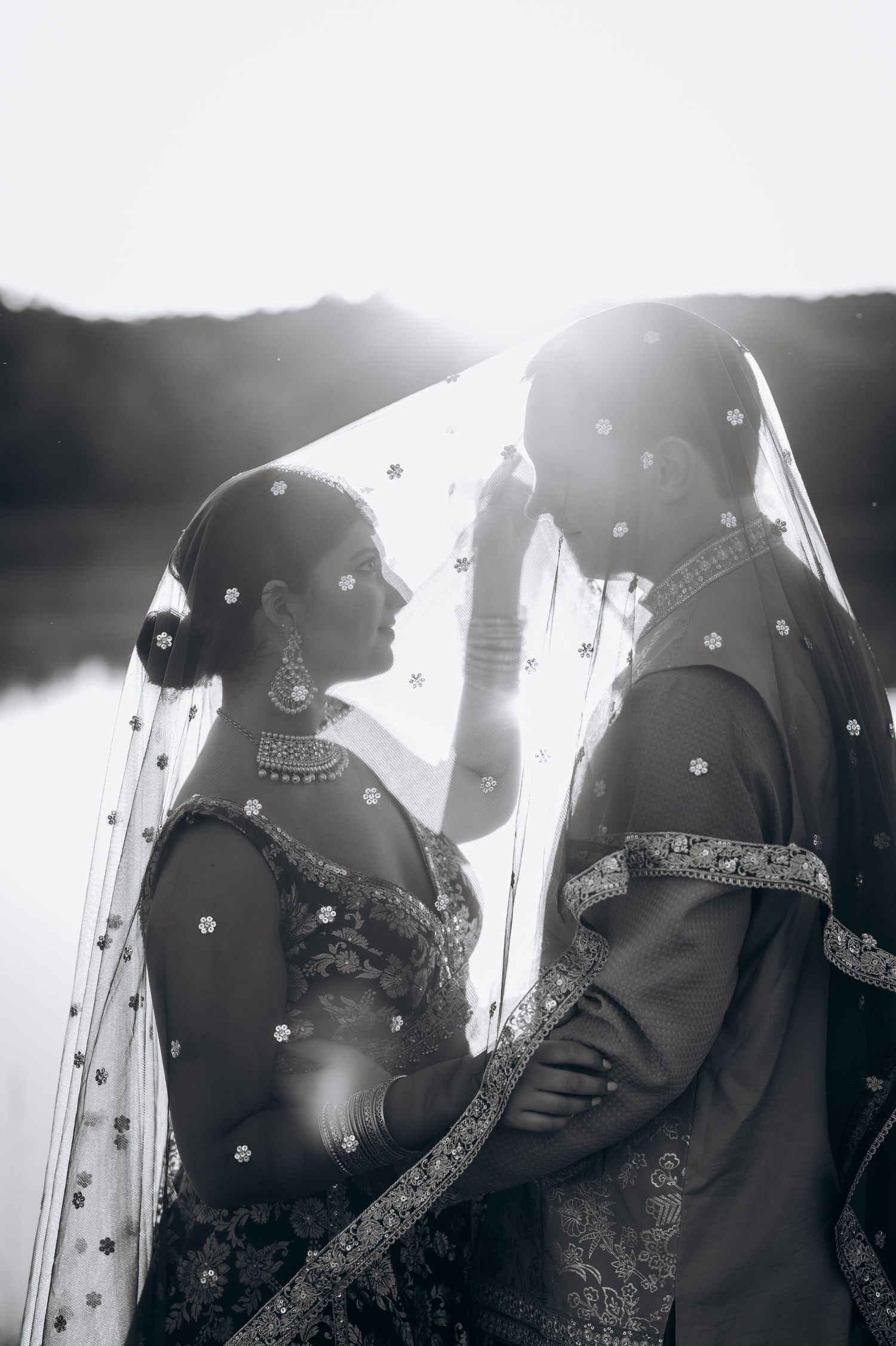 Black and white photo of Indian bride and groom sharing intimate moment under embroidered veil with sunlight backdrop.