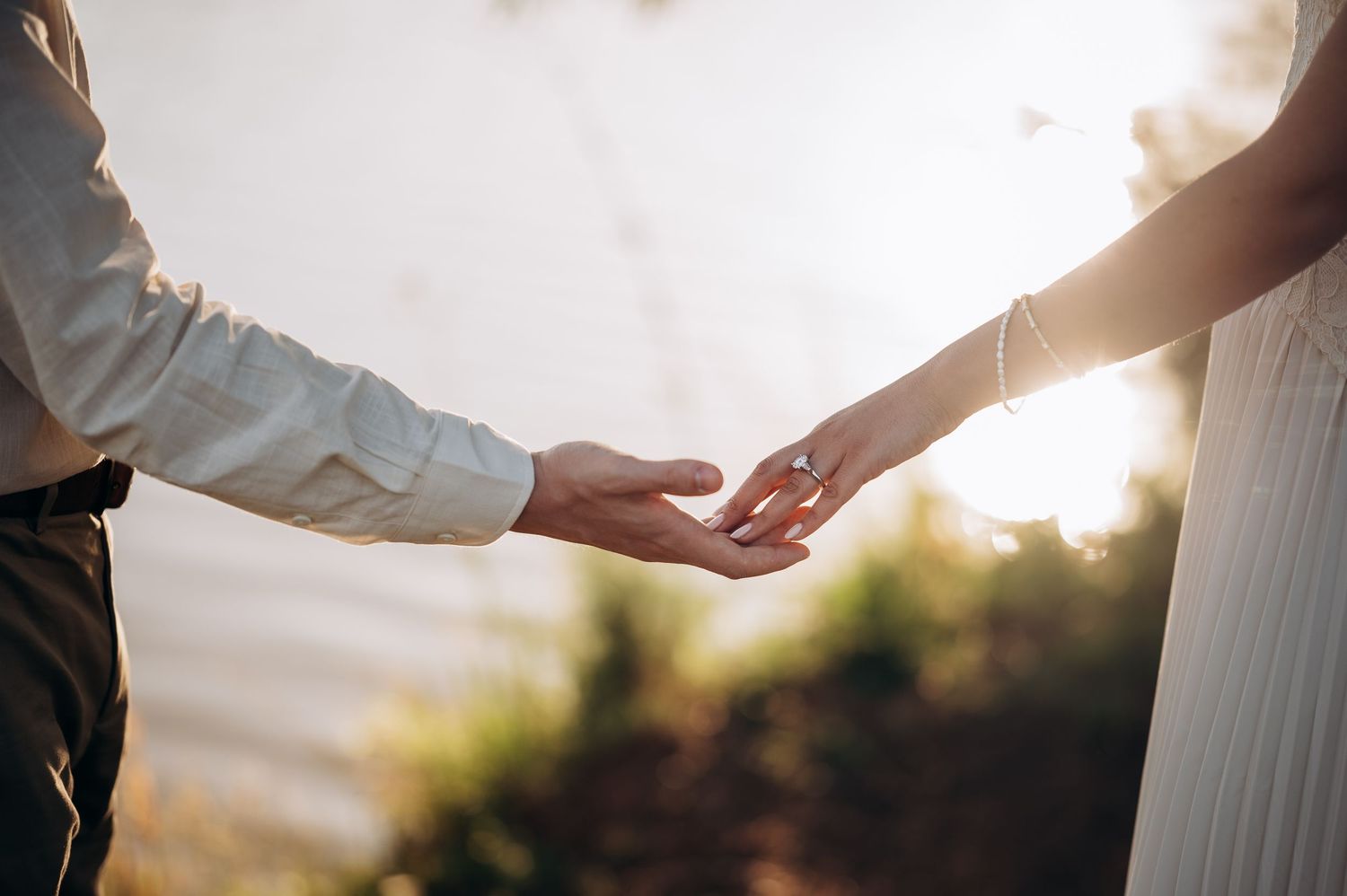 Couple holding hands outdoors at sunset, woman wearing engagement ring, romantic golden hour lighting.