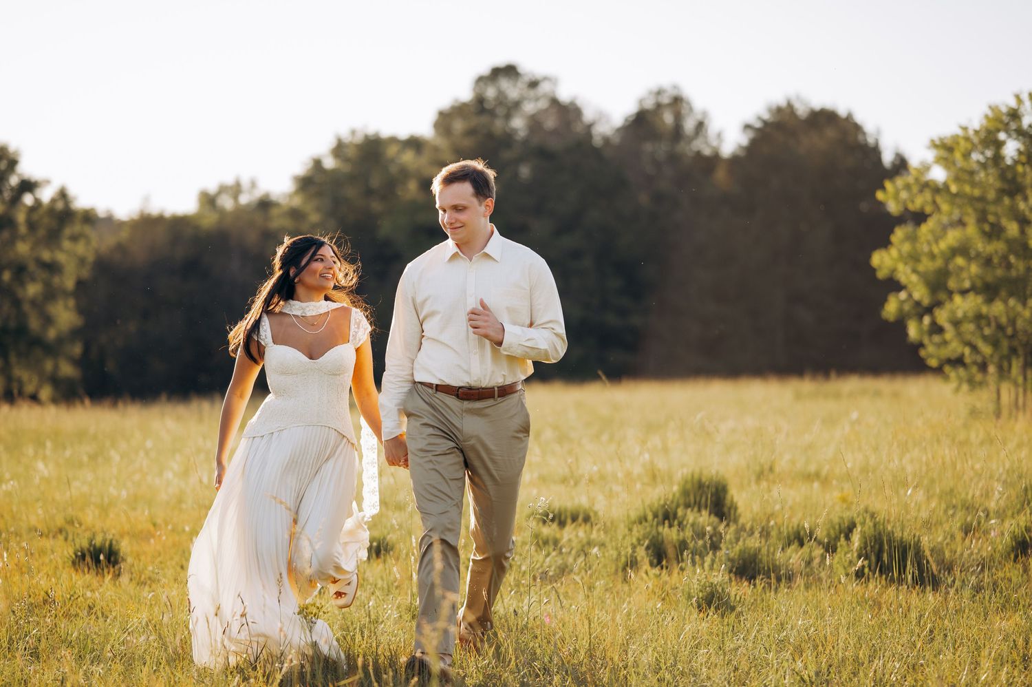 Smiling bride in white gown and groom in khakis walk together through a beautiful sunlit grassy field.