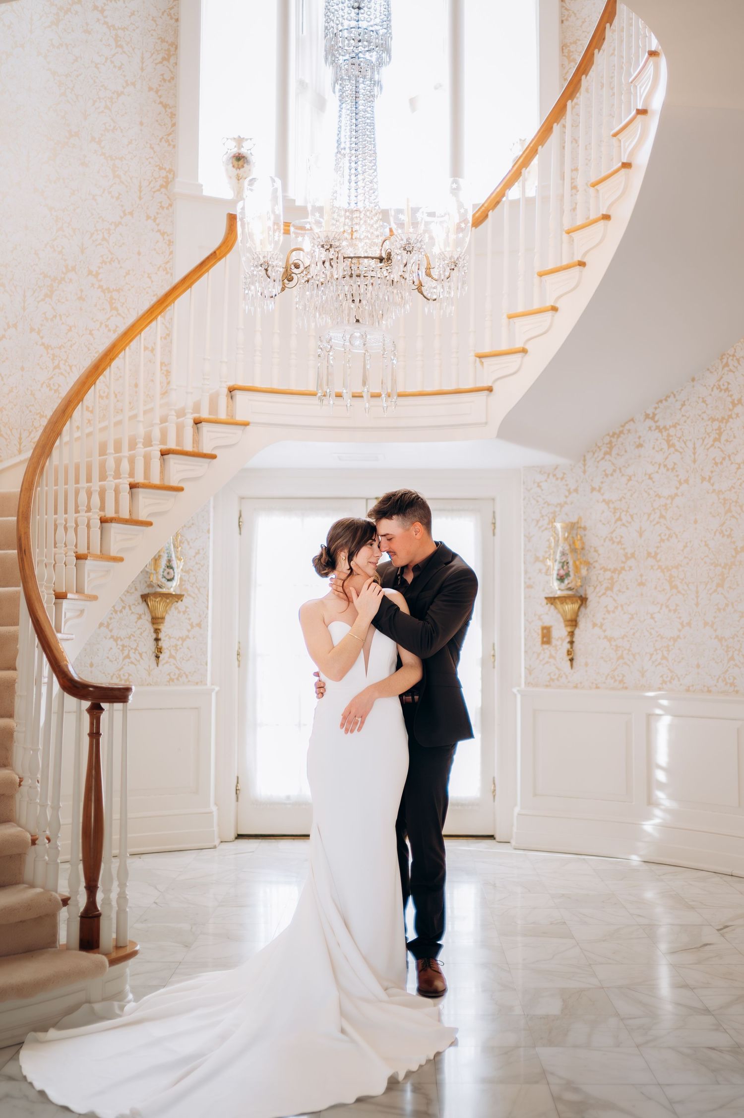 Bride and groom embrace in elegant mansion foyer with grand curved staircase and crystal chandelier.