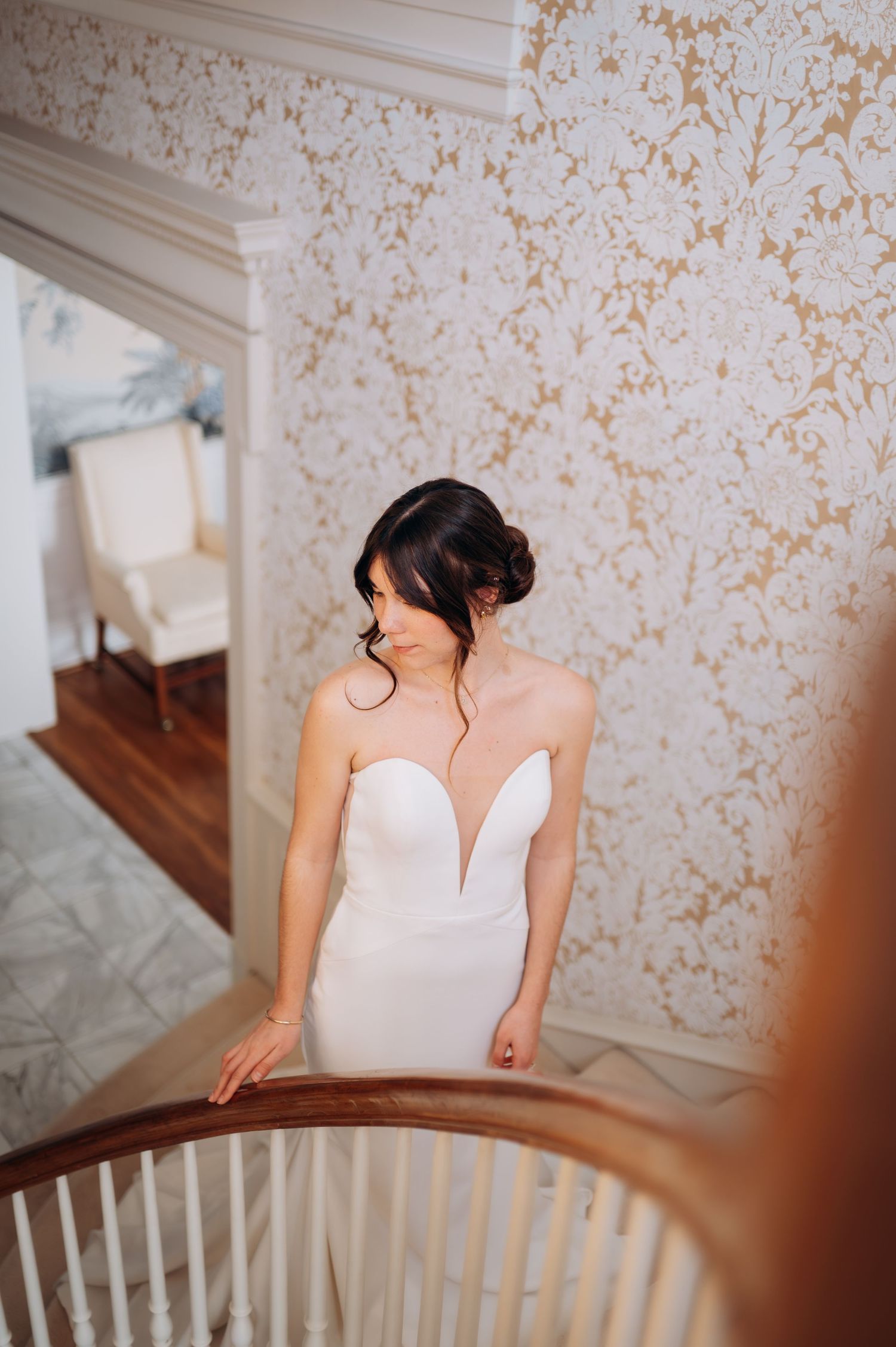 Bride in strapless white plunging neckline wedding gown poses on elegant staircase with gold floral wallpaper.