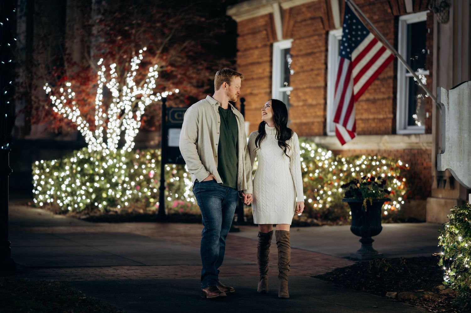 Couple holding hands and gazing at each other near a home decorated with Christmas lights and flag.