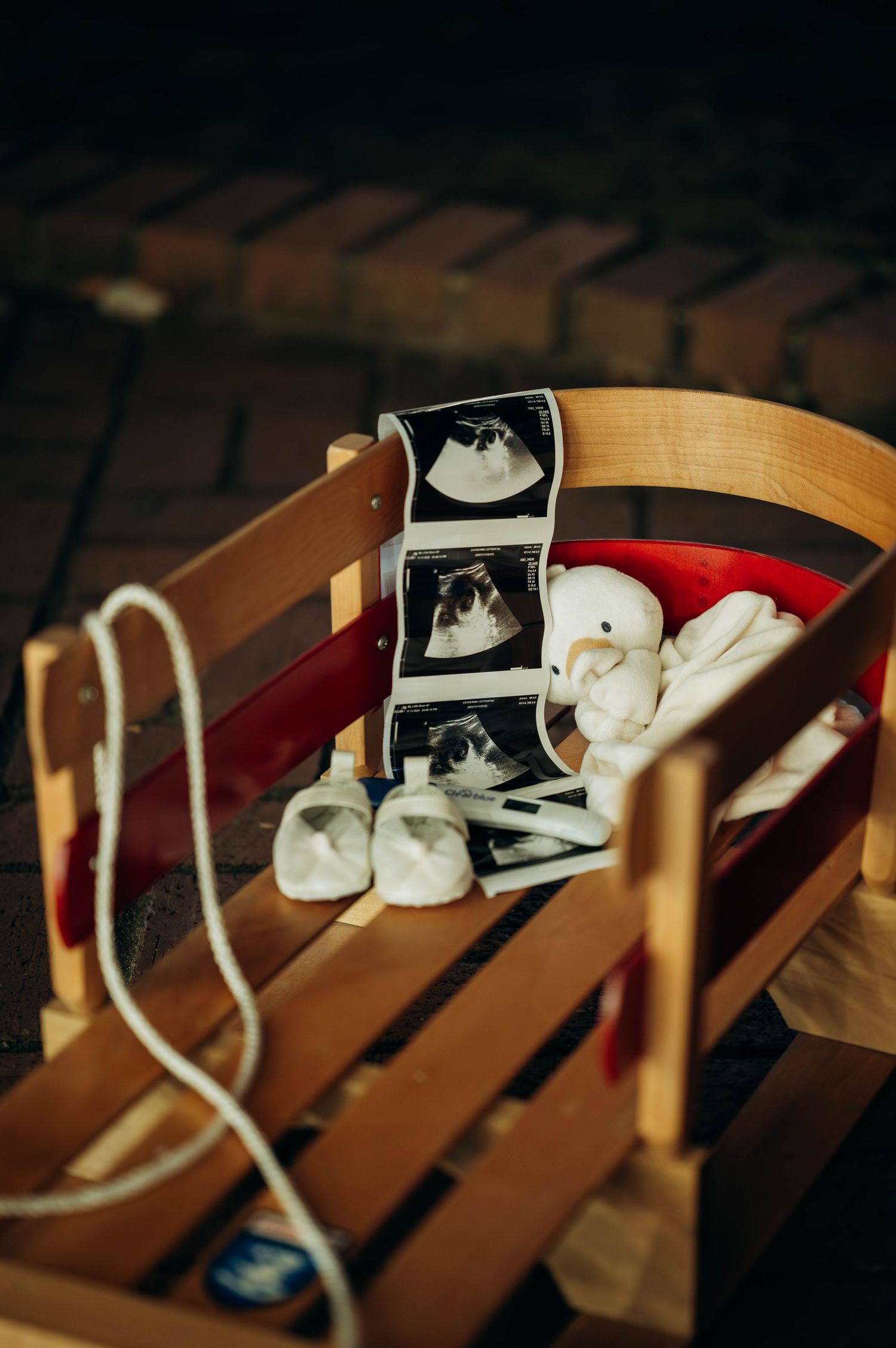 Wooden sled holding ultrasound photos and tiny white baby booties on brick pavement, pregnancy announcement keepsake.