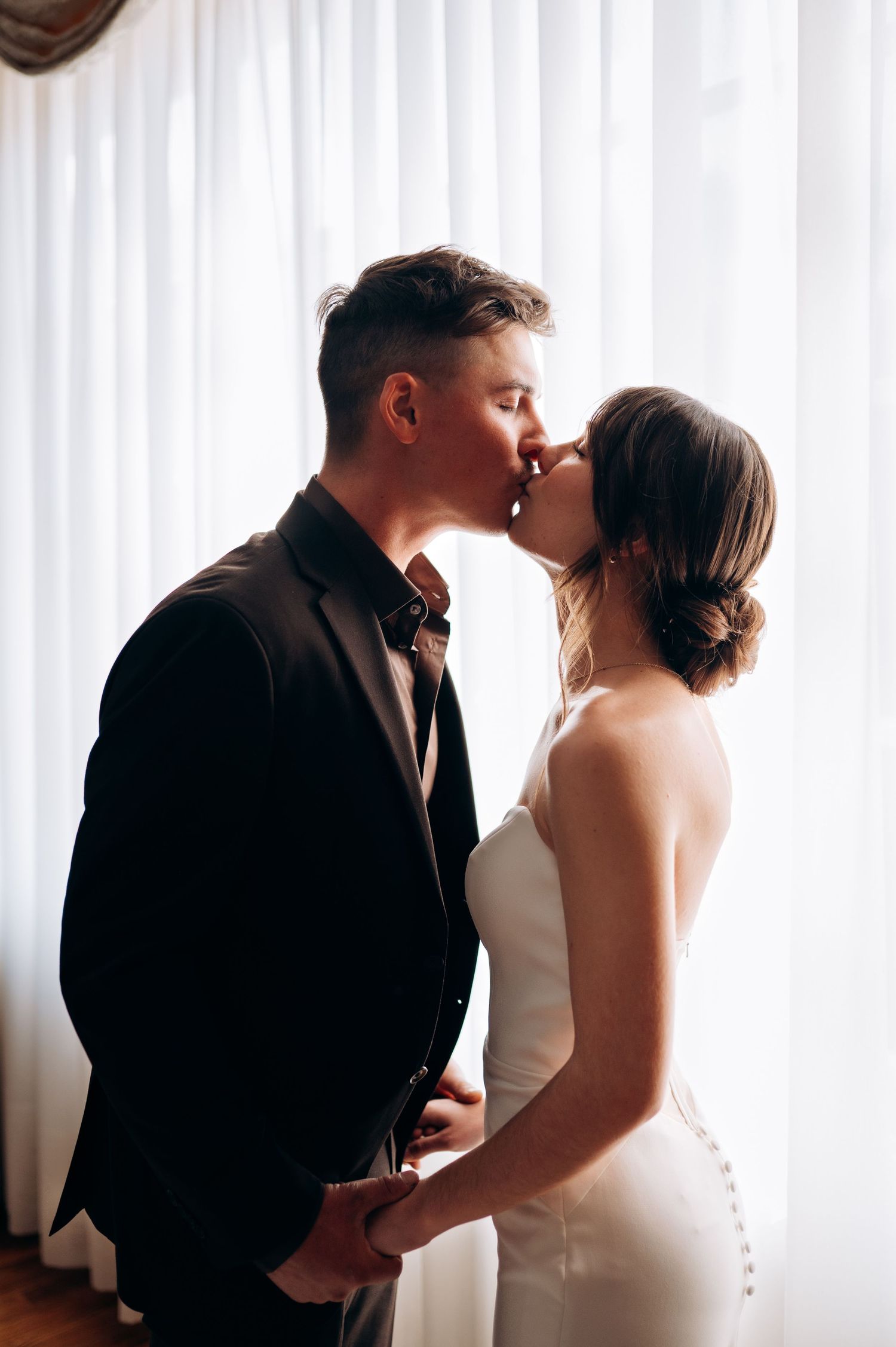 Bride and groom sharing romantic kiss silhouetted against bright white curtained window.