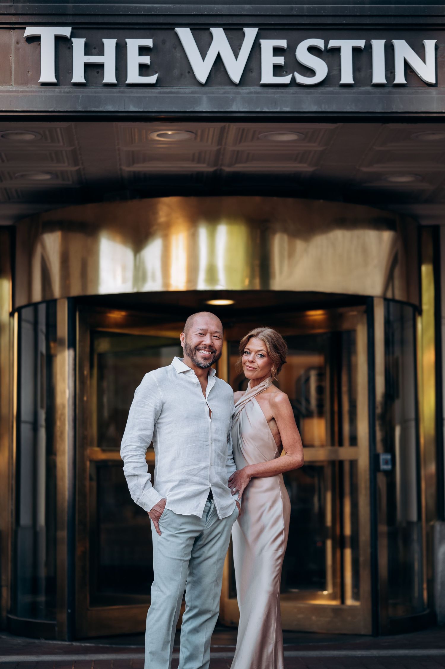 Elegant couple posing in front of The Westin Hotel entrance, man in white shirt and woman in champagne gown.