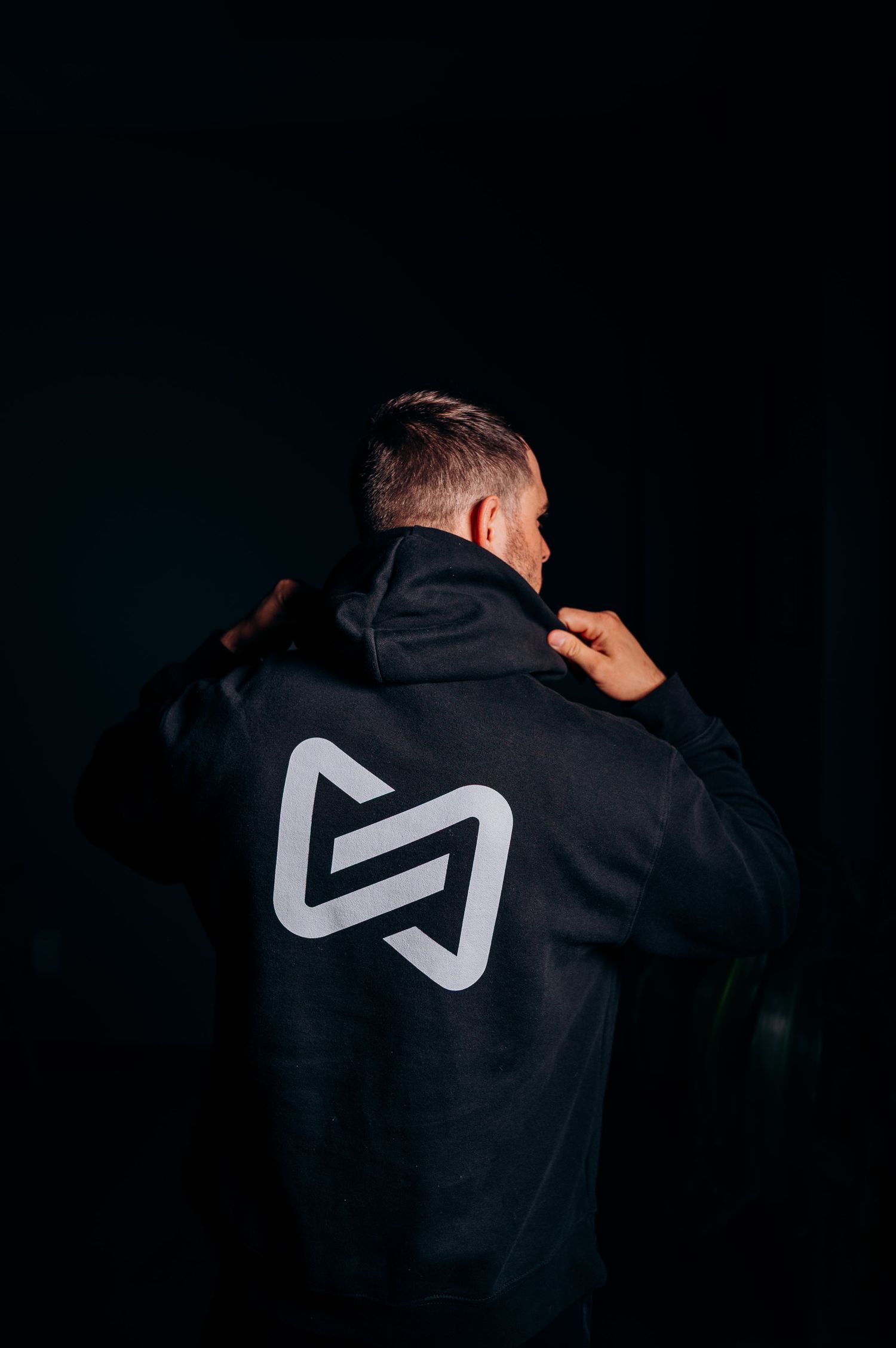 Person wearing a black hoodie with geometric logo design photographed from behind in dark lighting.
