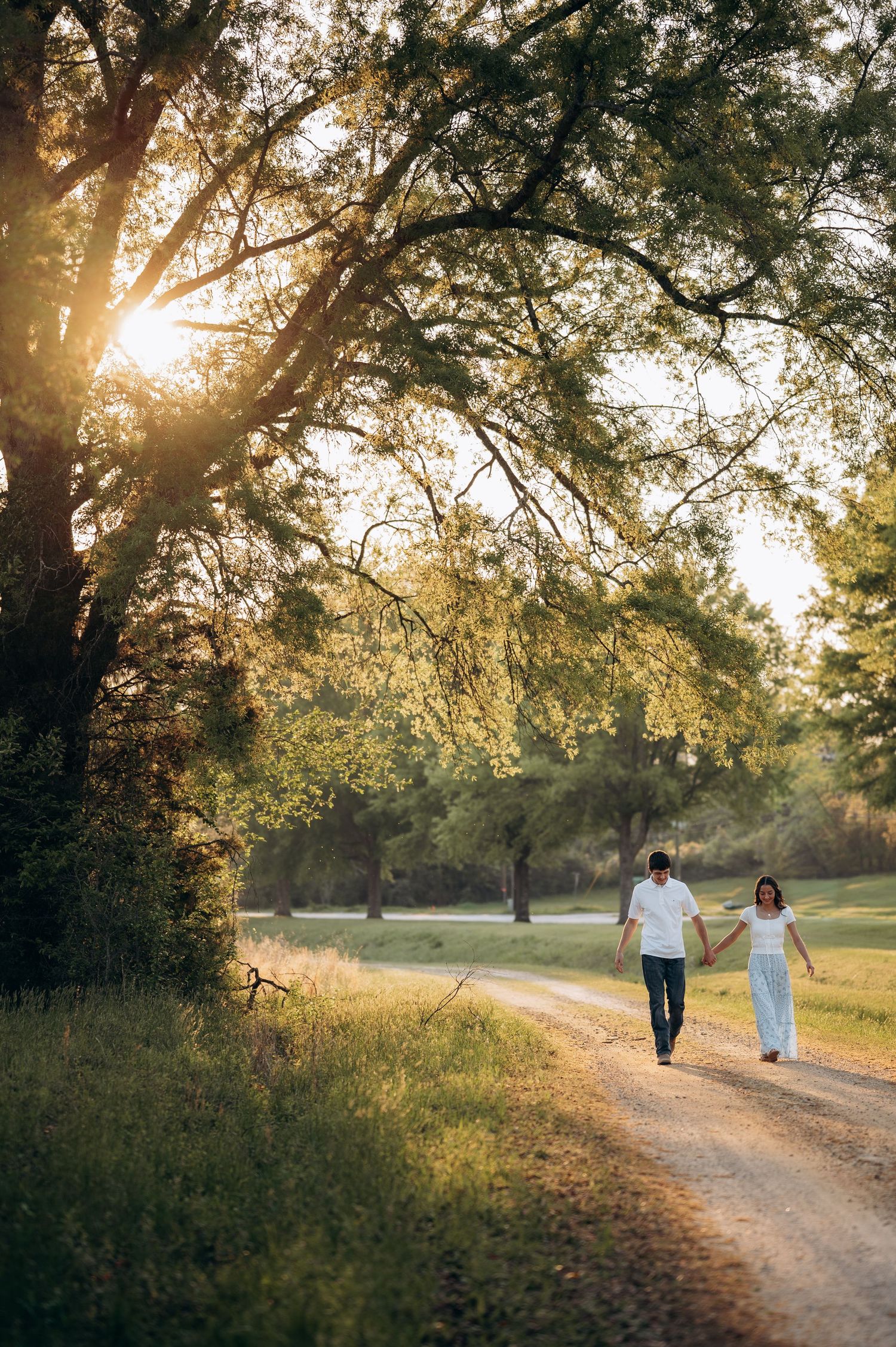 Couple holding hands walking on a sunlit dirt path surrounded by lush green trees at golden hour.