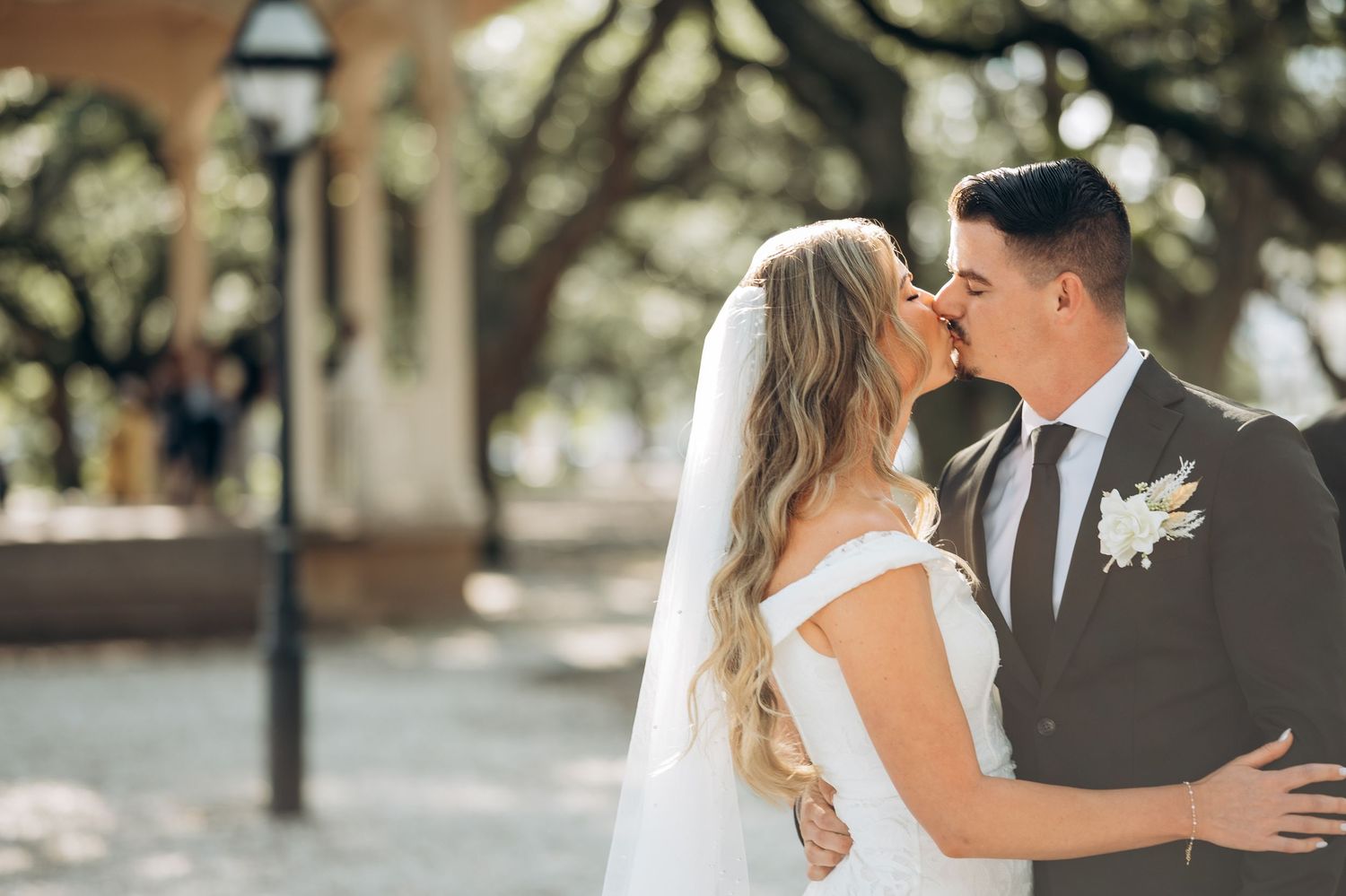 A bride and groom share a romantic kiss outdoors, surrounded by lush trees, as she wears a white off-shoulder gown.