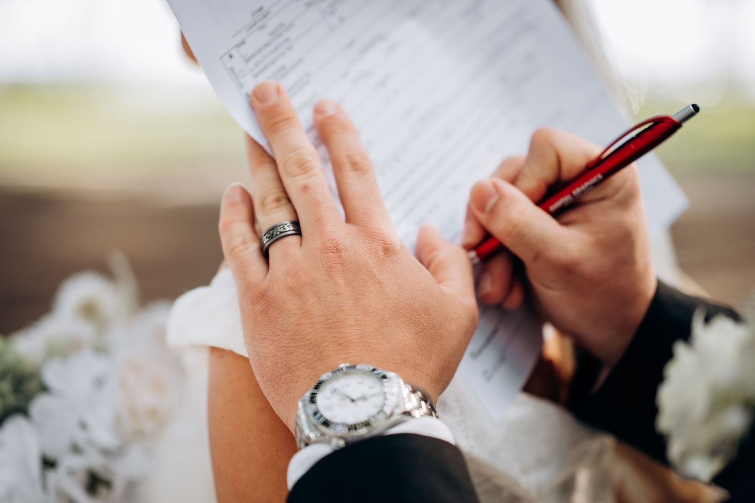 Close-up of groom signing wedding certificate with bride's hand resting on his.