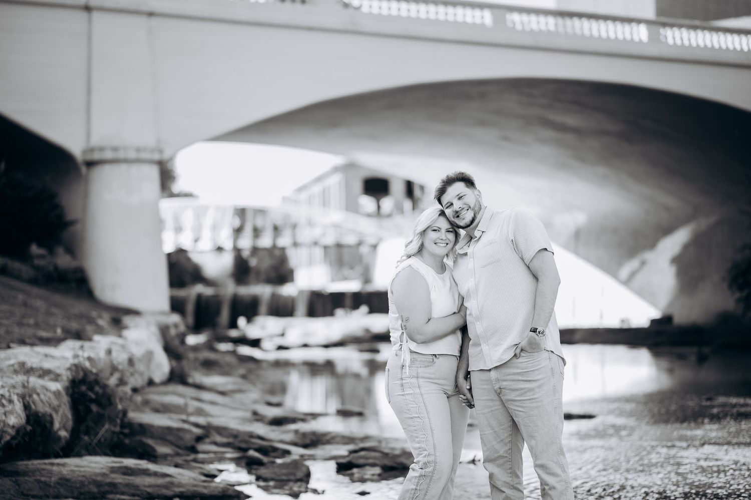 Black and white photo of a couple embracing and smiling under a stone bridge near a rocky waterway.