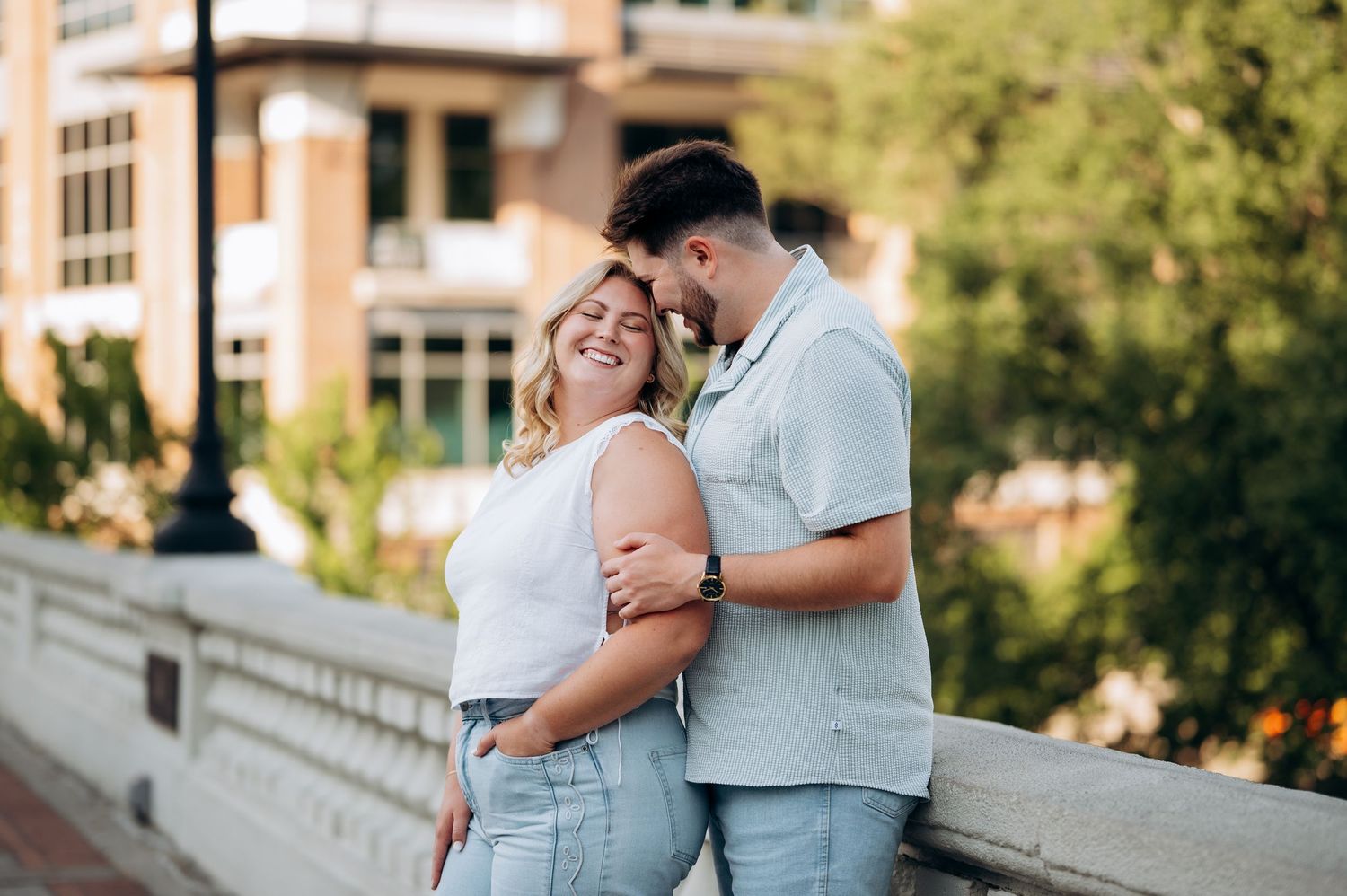 A smiling couple embraces on a bridge in an urban setting, the man nuzzling the woman's forehead affectionately.