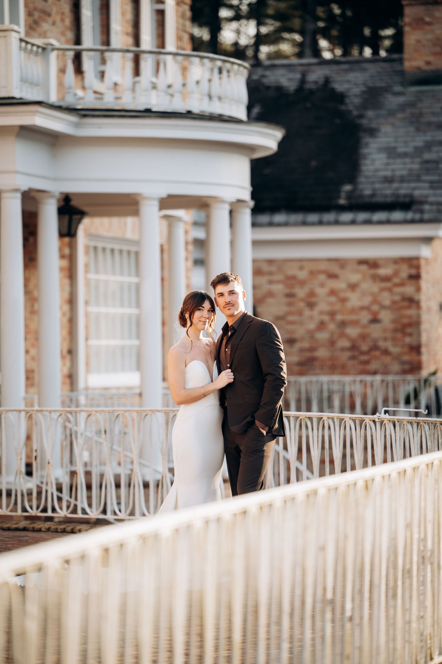 Elegant bride and groom posing on a grand estate balcony with white columns and ornate railings on their wedding day.