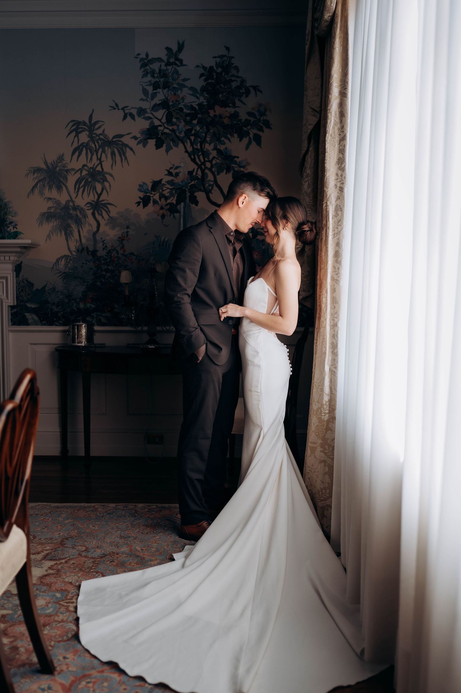 Elegant bride in a fitted white gown and groom in a dark suit share an intimate moment by a window.